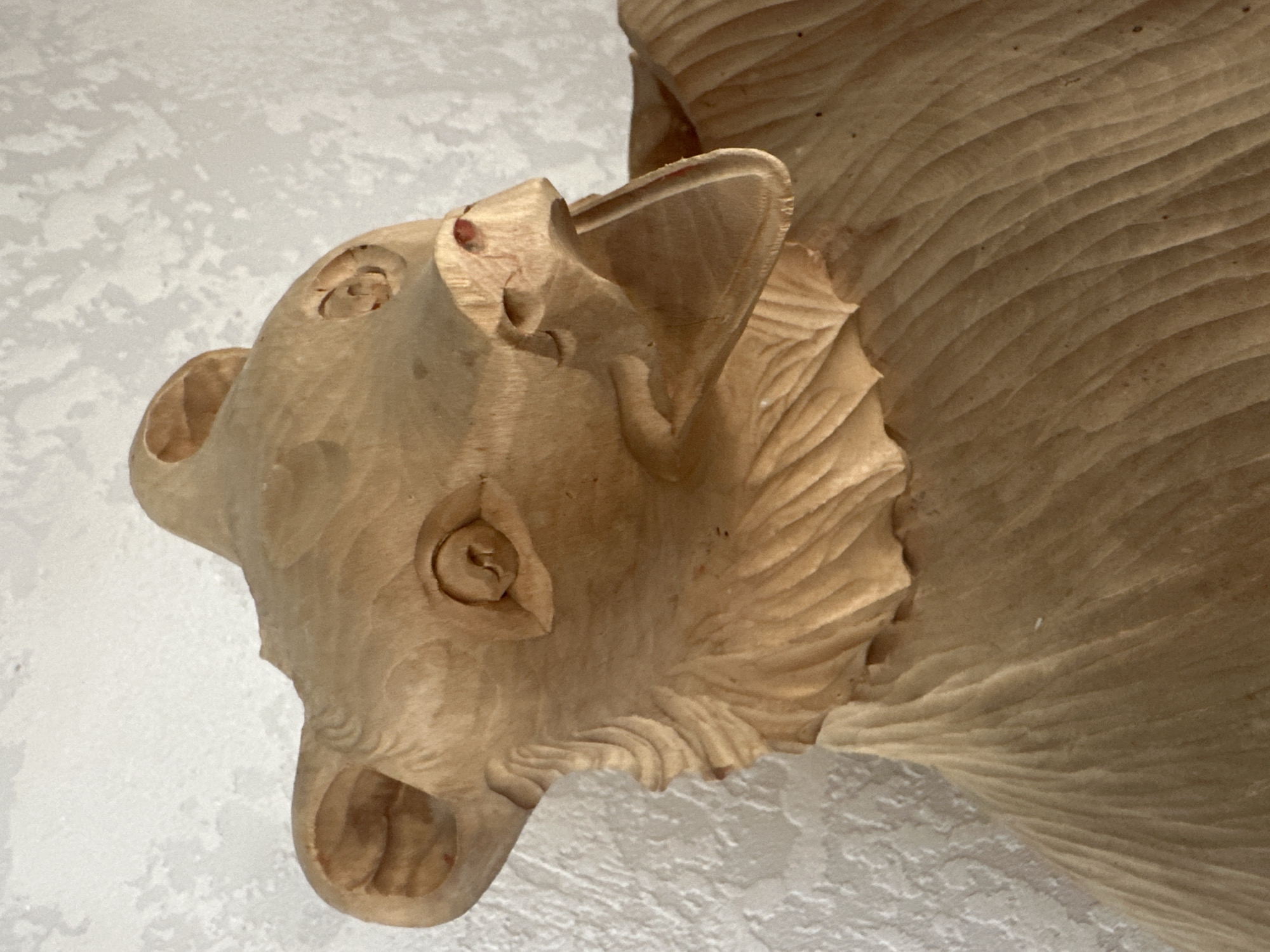 A close-up, angled shot shows a wooden carving of a bear's head and upper body. The carving is made of light-colored wood and displays detailed textures, particularly in the fur around the neck and the grain of the wood on the body. The bear's mouth is open, revealing the inside of its mouth and a hint of red on its snout. The background is a textured, off-white surface, likely a wall. There is no visible context to suggest a specific location in Yessentuki, Russia.