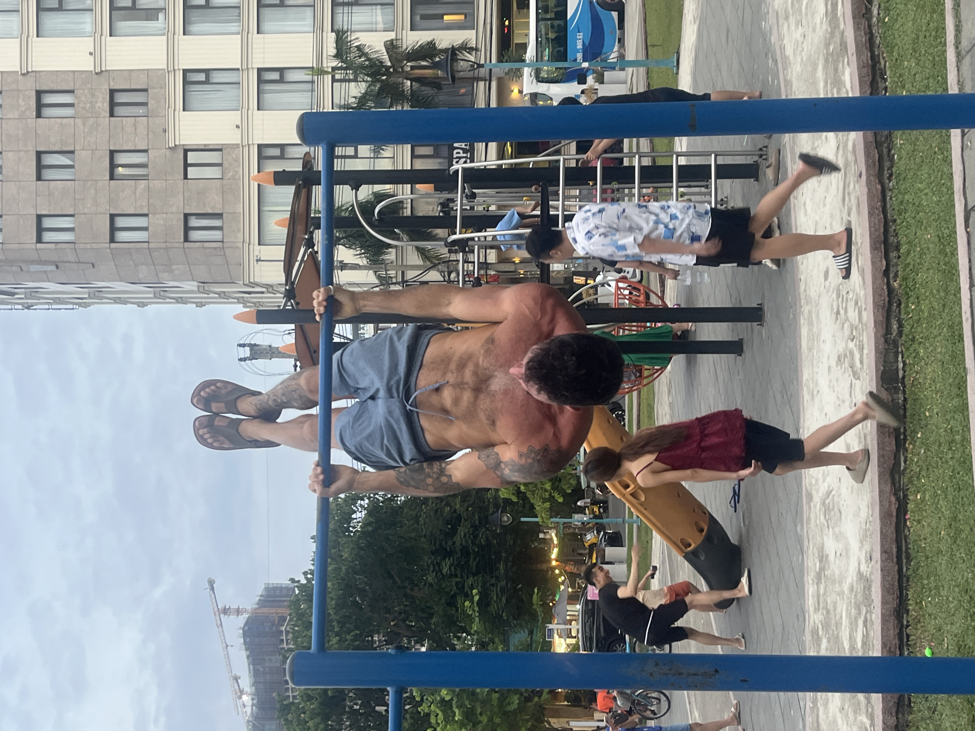 In Da Nang, Vietnam, a man is performing an inversion exercise on a pull-up bar in an outdoor public fitness area. He is holding the bar with his hands, with his body suspended upside down and his legs bent. He is wearing blue shorts and flip-flops. Several other people are in the vicinity. In the background, a large building with multiple windows is visible, along with trees and a bus. Construction cranes are also present in the distance. The ground is paved, with grass areas surrounding the fitness equipment.