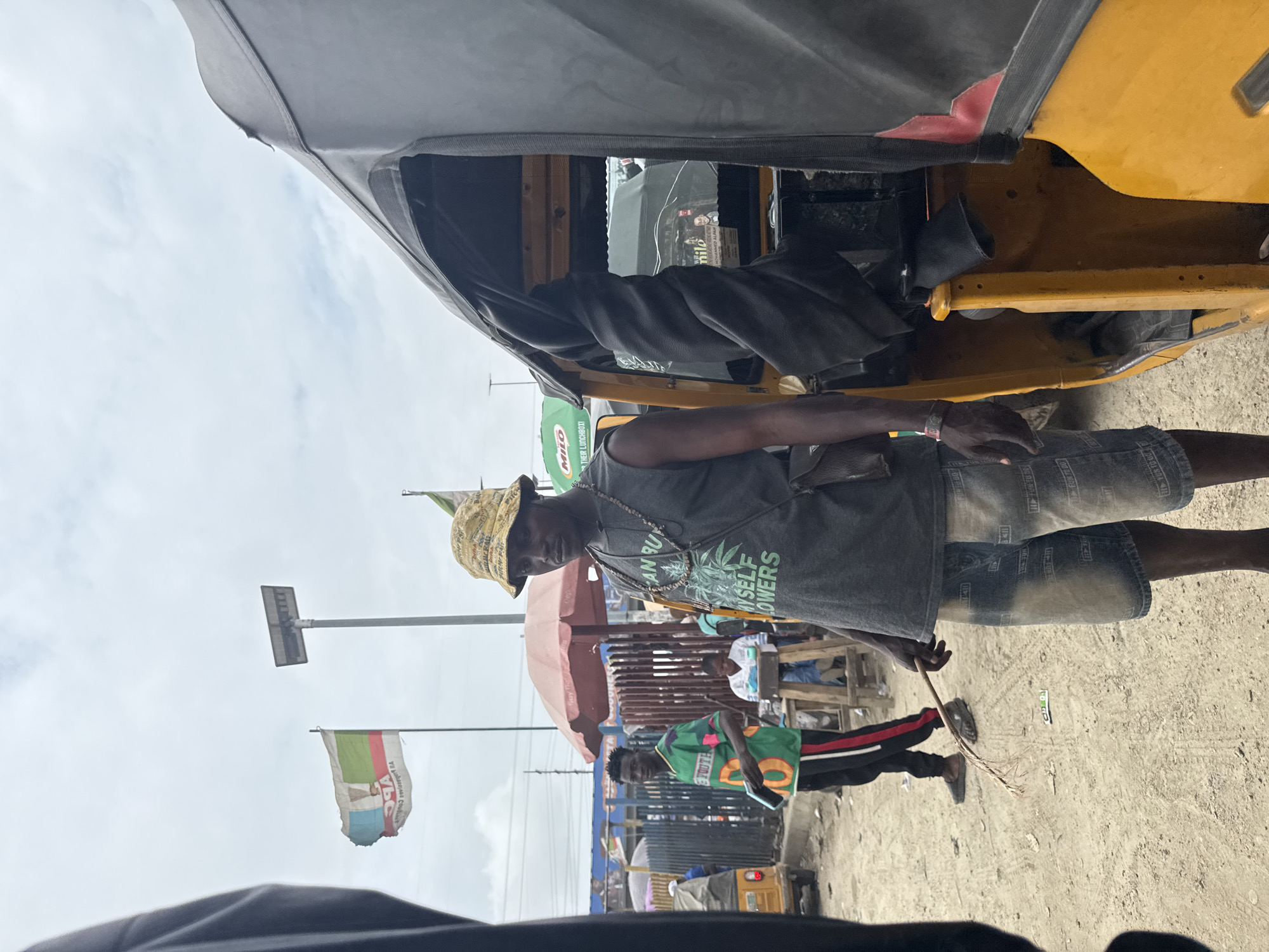 A man wearing a bucket hat, a gray sleeveless shirt, and denim shorts stands in front of a yellow and black auto-rickshaw. He holds a broom made of twigs in his right hand. In the background, two other men are visible, one in a green t-shirt and the other in a white shirt. Two flags, one with green, white, and red sections and another with green and white, are planted in the background. The scene appears to be a street in Lekki, Nigeria, with a cloudy sky overhead.