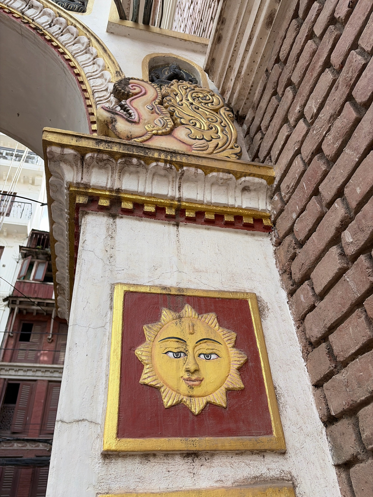 The image shows a close-up view of an architectural detail in Lalitpur, Nepal. The primary subject is a section of a white-plastered column adorned with decorative elements. At the lower half of the column, there is a rectangular panel with a gold border surrounding a deep red background. Centered within this panel is a golden relief of a sun with a human face. The sun has rays emanating outwards, and the face is depicted with distinct eyes, nose, and lips. The top section of the column features ornate plasterwork and a golden decorative motif, possibly a stylized animal head, protruding from an archway. To the right of the column, a textured red brick wall is visible, suggesting the building's construction. In the background, out of focus, are parts of other buildings, one appearing to be a multi-story structure with dark red wooden shutters and balconies. The overall impression is of traditional architecture, likely a temple or heritage building, showcasing intricate craftsmanship.