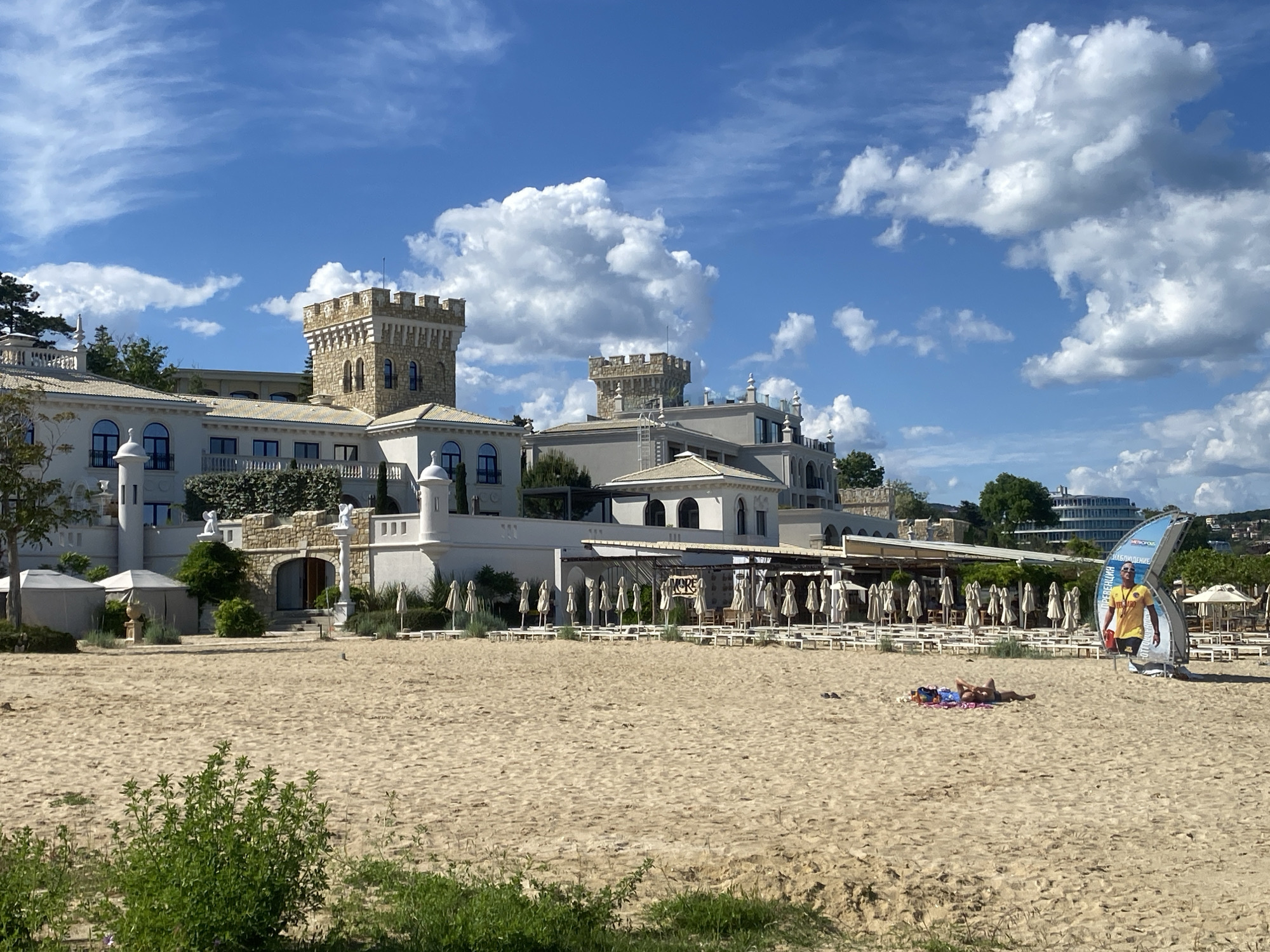 The image depicts a sunny beach in Varna, Bulgaria, with a distinctive castle-like resort building in the background. The building is off-white, with ornate towers and a section with arched openings leading to a beachfront terrace. On the beach, there are rows of beach chairs and umbrellas, suggesting a resort or private beach area. Two people are lying on the sand sunbathing. A lifeguard advertisement is placed on the sand near the couple. The sand is light tan, and there is vegetation in the foreground, partially obscuring the view of the beach. The sky is blue with scattered puffy clouds, indicating a pleasant weather. A modern building is also visible in the far background.