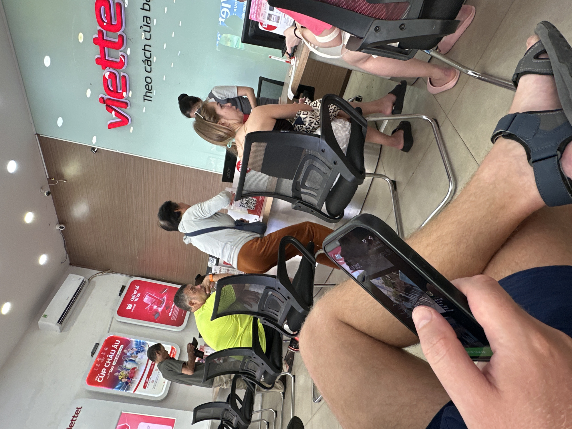 The image depicts an indoor waiting area of a telecommunications service center, likely Viettel, located in Nha Trang Ward, Vietnam. The photograph is taken from a low angle, looking across the room, and is originally rotated counter-clockwise.
In the immediate foreground, the lower half of a person is visible, wearing blue shorts and dark blue sandals. Their hands are holding a black smartphone, which displays text including "Latest Staked" on its screen.
Several other individuals are seated in the room, appearing to be waiting or being assisted. A man in a bright yellow shirt is seated, facing away from the camera, with another man in a grey shirt further to the left. Further back, a woman with blonde hair in a dark top is seated at a desk, looking towards a counter. Another individual in a white shirt and brown pants is also seated, facing towards the right.
The background features a light green wall with a prominent, red 3D "Viettel" logo and the Vietnamese slogan "Theo cách của bạn" (meaning "Your way"). Below this, there's a section of wooden paneling. Mounted on the wall are several illuminated display signs. Visible text on these signs includes "CÚP CHÂU ÂU" (Europe Cup), and "CHỮ KÝ SỐ TỪ XA" (Remote Digital Signature) with "mysign" and "MIỄN PHÍ DỊCH VỤ" (Free Service). An "acer" logo is visible on a monitor screen. An air conditioning unit is installed high on the left wall. The general lighting suggests it is daytime.