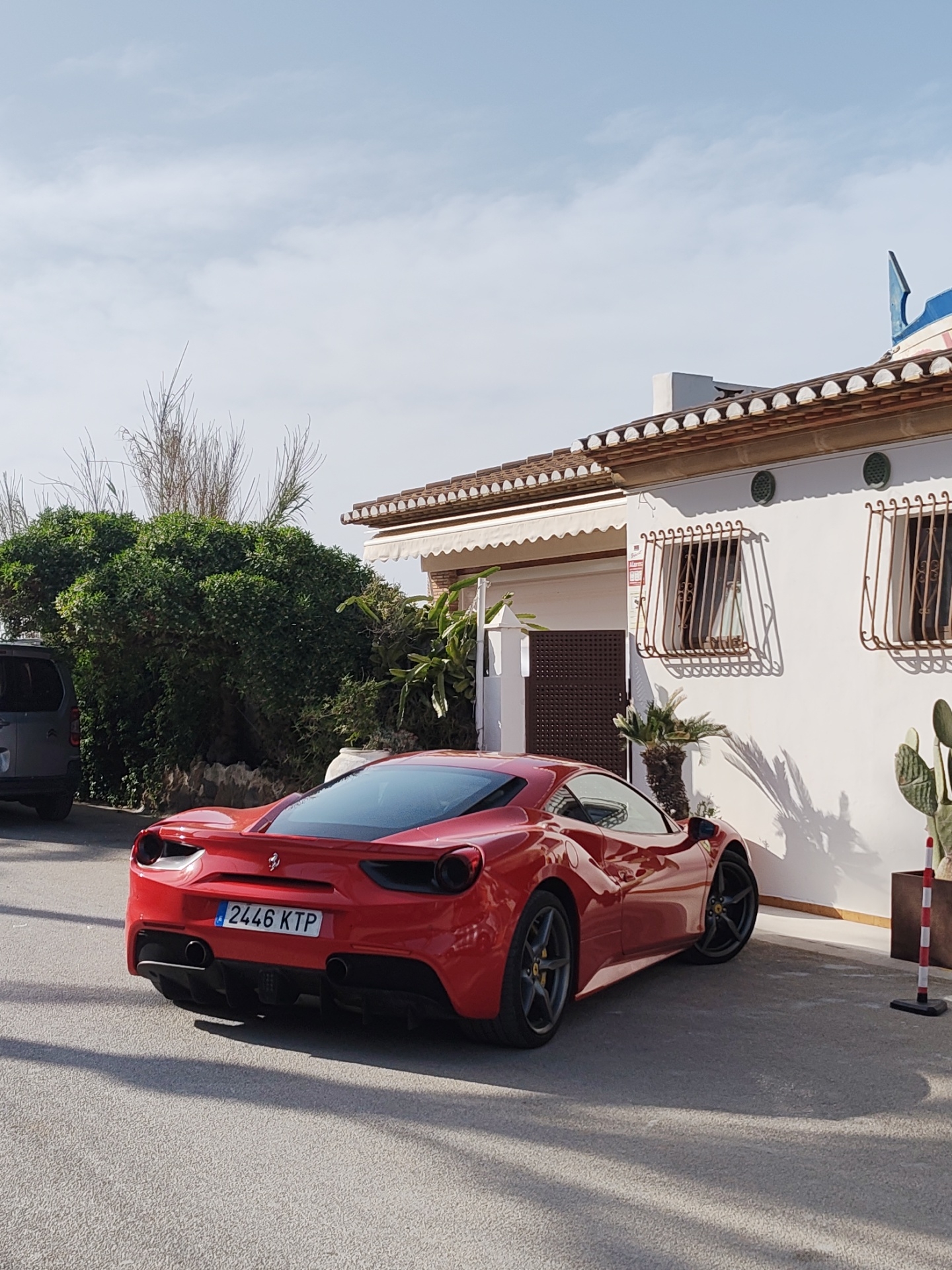 A red Ferrari 488 Spider is parked on a paved surface in front of a white building in Dénia, Spain. The car's license plate reads "2446 KTP." The building has two barred windows, a white awning, and a tiled roof. A large green bush is to the left of the building, and a grey vehicle is partially visible behind the bush. A potted cactus is next to the building's entrance. The sky is partly cloudy.