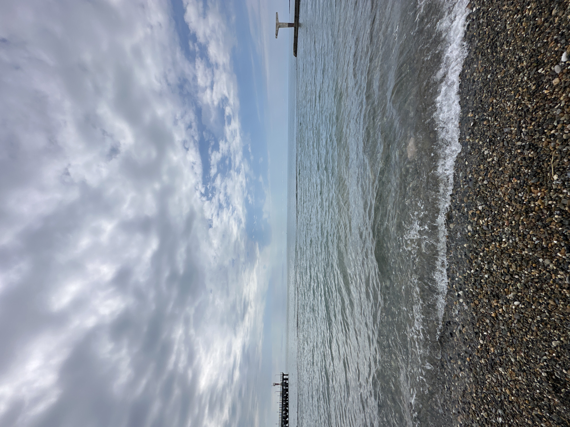This image depicts a serene coastal landscape, likely in Sochi, Russia, captured during daylight hours under a partially cloudy sky. The photograph is taken from an eye-level perspective at the shoreline, looking out towards the sea and horizon.
The upper portion of the frame is dominated by the sky, filled with a mix of soft white and light grey clouds. Patches of blue sky are visible through the cloud cover, particularly towards the upper right. The clouds exhibit various forms, from wispy streaks to more substantial, textured masses.
A distinct horizon line separates the sky from the vast expanse of the sea in the middle of the image. The water appears calm, with subtle ripples and gentle waves visible across its surface, reflecting the overcast light. Two dark, man-made structures are noticeable along the horizon. On the left side, a low-lying linear structure extends into the water. Further to the right, a more prominent, T-shaped structure rises from the sea, resembling a section of a pier or breakwater.
The lower part of the image showcases a pebbly beach. The shoreline is composed of numerous small to medium-sized stones and pebbles, predominantly in dark grey, brown, and black tones. Gentle waves are breaking onto the shore, creating a thin line of white foam where the water meets the land. The water close to the shore is relatively clear, allowing some of the submerged pebbles to be seen.
There are no people or discernible text visible in the photograph. The scene conveys a tranquil and natural ambiance.