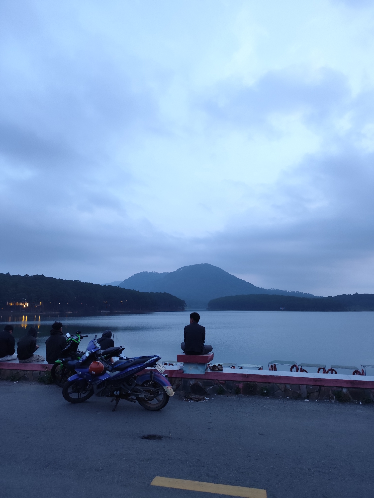 The image shows a view of Xuan Huong Lake in Da Lat, Vietnam, under a cloudy sky. In the foreground, there's a road with a yellow painted stripe and two motorcycles parked along the side. People are sitting along the edge of a low wall that separates the road from the lake, some looking towards the water. One individual is sitting cross-legged on the wall, facing the lake. In the middle distance, there is a large body of water, with forested hills and mountains visible in the background. A series of small white boats is lined up along the water’s edge near the wall. The overall scene is captured in a slightly dim, blueish light.