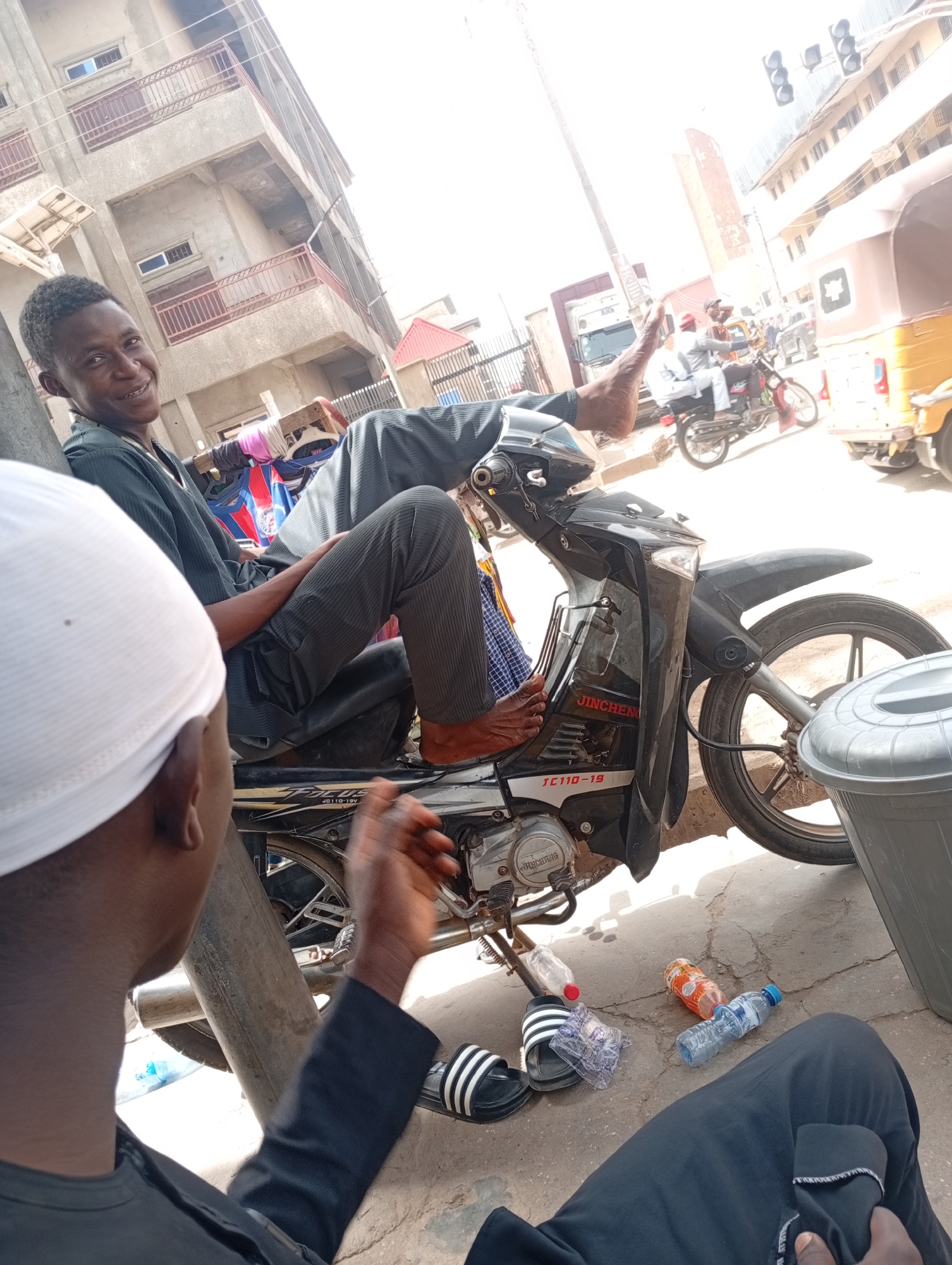 Two young men are in an outdoor scene, likely Maiburiji, Nigeria. One man is seated on a black Jincheng motorcycle with registration JC110-19. He is smiling, wearing a dark shirt and striped pants, and his feet are bare. The other man, wearing a white cap and dark shirt, is partly visible. There are water bottles and sandals on the ground near the motorcycle. In the background, there is a building, other motorcycles and vehicles, and traffic lights.