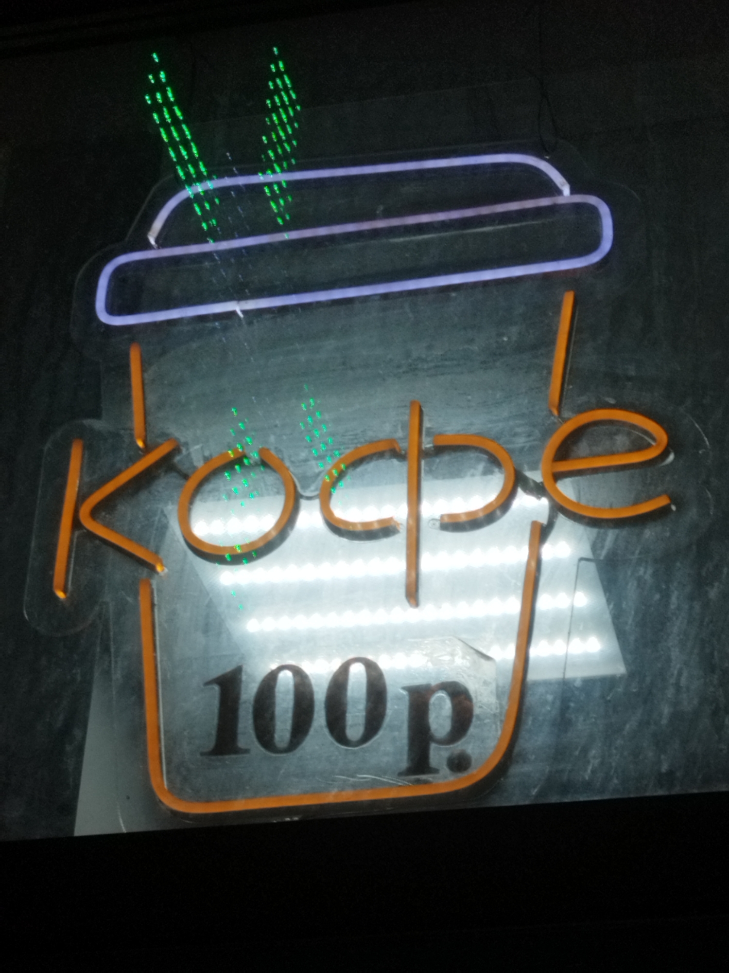 The image shows a neon sign for a coffee shop in Surgut, Russia. The sign is shaped like a coffee cup with steam rising from it. The cup is orange, and the steam is green. The words "кофе" (coffee) and "100 р." (100 rubles) are written on the cup in orange and black, respectively. The sign is illuminated by a bright white light. The background is dark and appears to be a wall or a window. The image is taken at an angle, and there are some reflections on the surface of the sign.