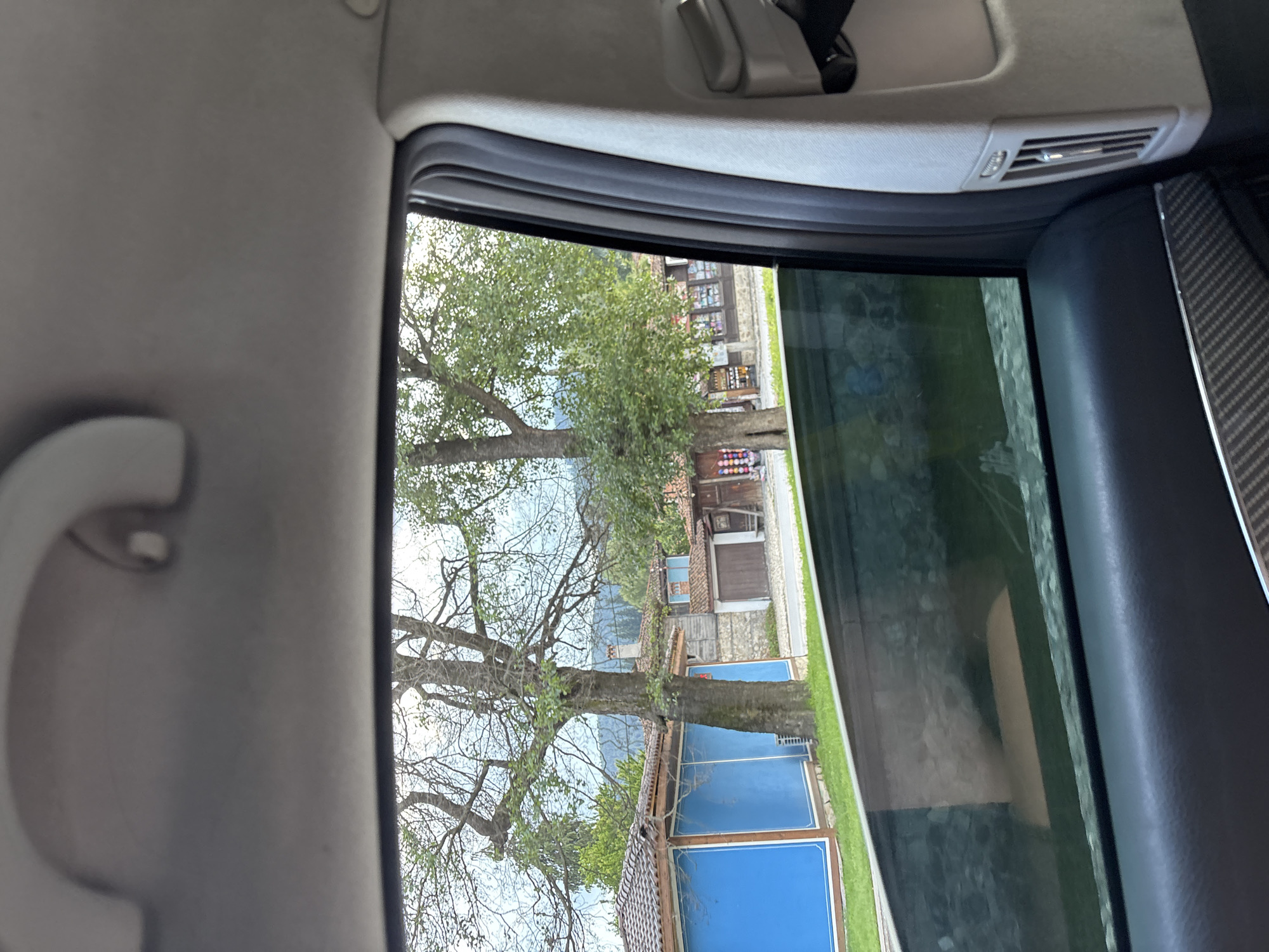 The image captures a view from inside a car, looking out through the side window. The scene outside depicts a village with traditional Bulgarian architecture, specifically in Koprivshtitsa. We see several rustic buildings with tiled roofs, some featuring wooden facades and stone elements. Bright blue rectangular structures, possibly modern additions or renovated sections, stand out against the older buildings.
A large, mature tree with bare branches dominates the foreground, its trunk thick and its limbs spreading wide, suggesting it might be early spring or late autumn. Beyond the tree and buildings, hills or mountains are faintly visible in the distance, shrouded in a light haze. The sky is overcast, hinting at a mild, possibly cloudy day.
The interior of the car is visible on the left and right edges of the frame. The left side shows part of the car's headliner and a handle. The right side shows the car's door panel with a textured carbon fiber-like trim.
The overall impression is one of a peaceful, rural setting, looking out from the comfort of a vehicle. There are no people clearly visible, and no specific activity is taking place. The lighting suggests it is daytime, but the overcast sky prevents strong shadows. The location context of Koprivshtitsa, Bulgaria, adds to the understanding of the architectural style observed.