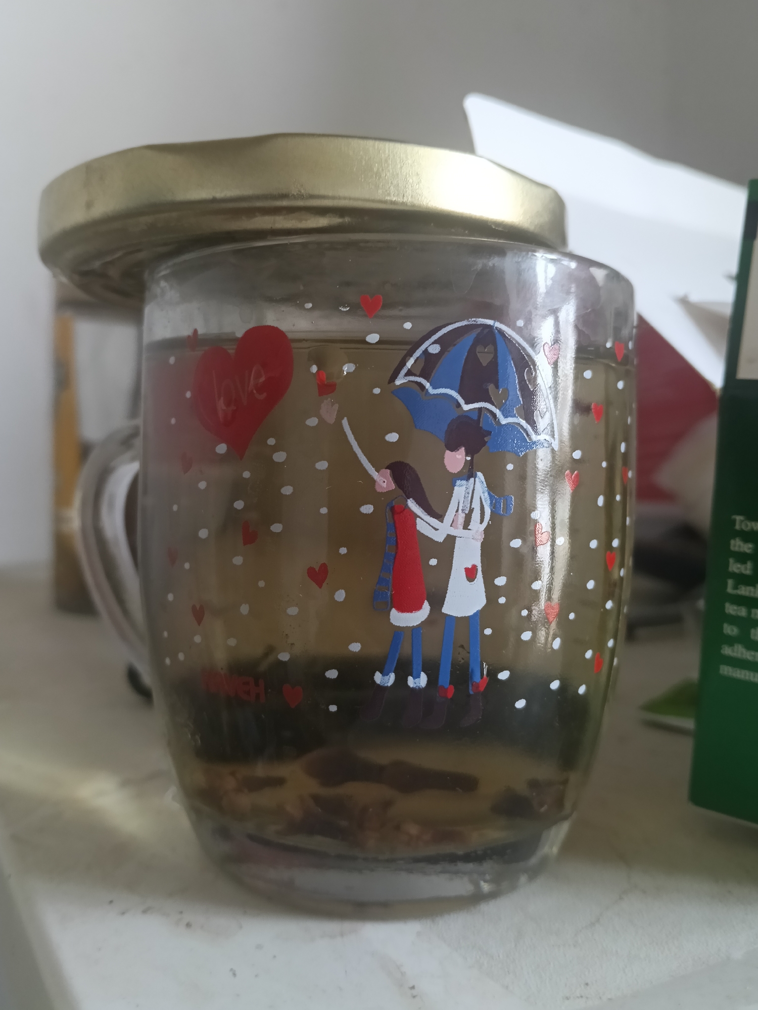 A close-up, eye-level shot shows a clear glass mug filled with a dark liquid, possibly tea, with visible tea leaves at the bottom. The mug is decorated with a pattern of red hearts and white dots. On the mug, there are two cartoon figures holding an umbrella, and a red heart with the word "love" inside. The mug is topped with a golden lid. In the background, a blurred white surface suggests a shelf or counter, with a white box and a green box with text on the right. The environment is an indoor setting, possibly a kitchen or pantry. The image is well-lit, with a slight focus on the mug. No location in Altinkul, Uzbekistan is discernable.