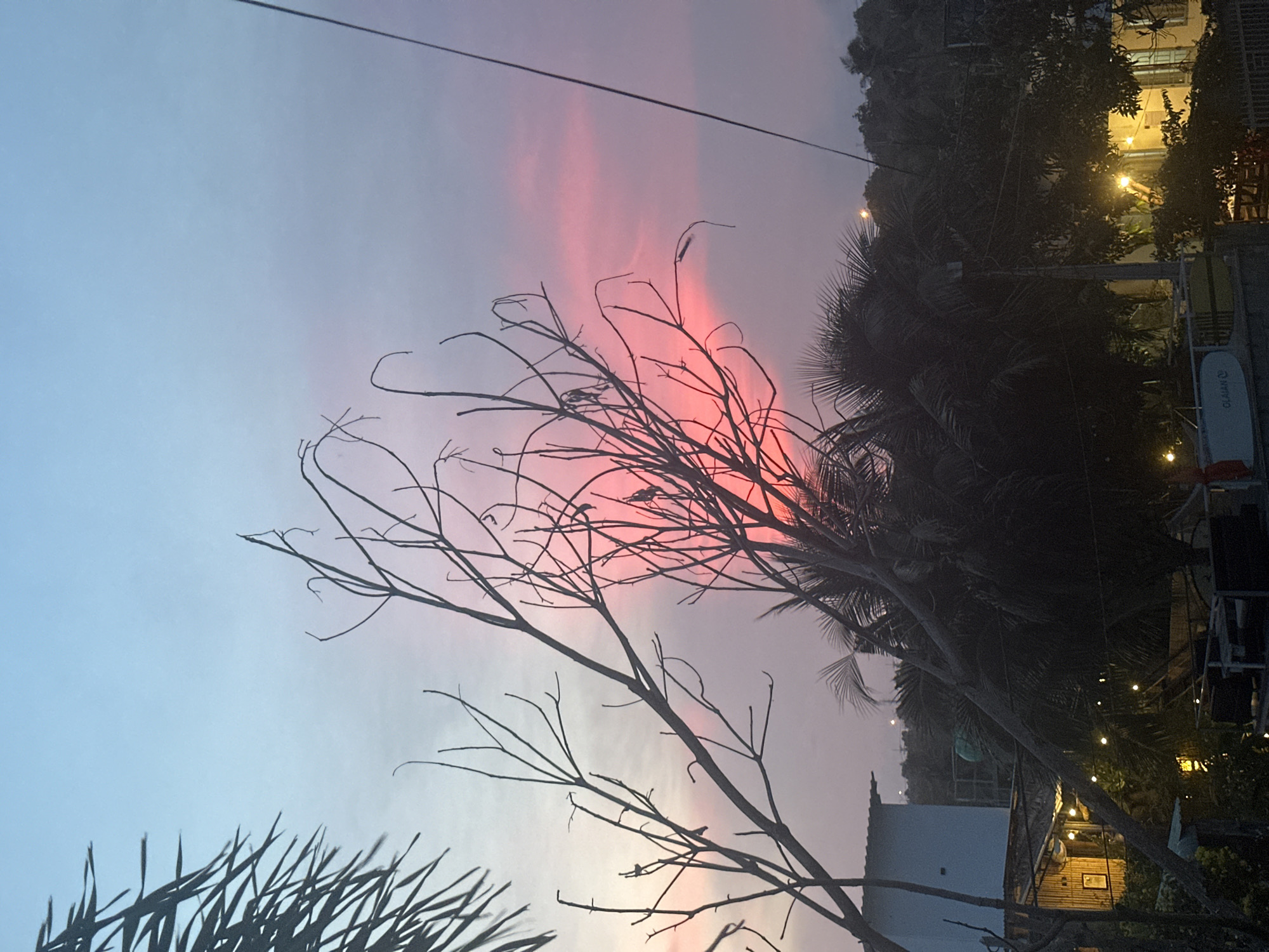 A sunset view over a tropical location. The sky displays a gradient of pale blue to pink hues. In the foreground, silhouetted tree branches, including palm fronds on the left, dominate the frame. In the midground, dark foliage of trees and palm trees are visible, interspersed with some buildings and lights. One building on the left is a light blue, two-story structure. Other structures appear to be bungalows or huts with illuminated string lights. In the background, a dark, hilly landscape is discernible against the sunset sky. A single black wire runs diagonally across the right side of the image. The overall scene depicts a serene, twilight atmosphere in a tropical environment.