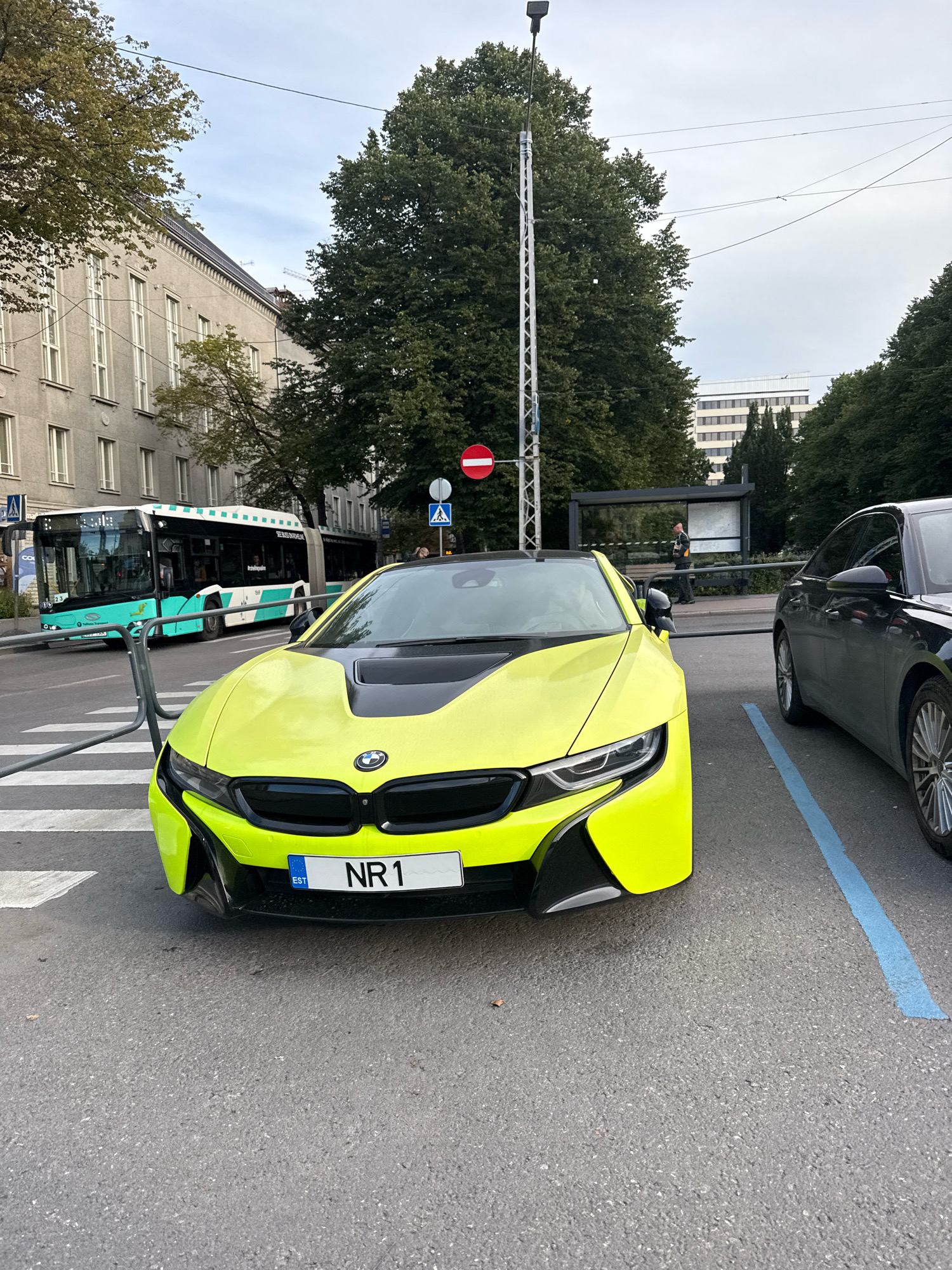 In Tallinn, Estonia, a bright yellow BMW i8 is parked on an asphalt street at a pedestrian crossing. The car has a black hood and black accents on its front bumper. Its license plate reads "NR1" with "EST" above it. To the right of the BMW, a dark grey sedan is parked, also on the asphalt. The scene includes a bus stop with a sign behind the BMW, and a bus, green and white, is visible on the street behind a metal barrier. Tall buildings with numerous windows line the street in the background, and large trees with green foliage are abundant. Overhead power lines are visible against a cloudy sky. One person can be seen standing at the bus stop, and another person is seated on the bus. A traffic sign indicating "no entry" is mounted on a pole near the bus stop.
