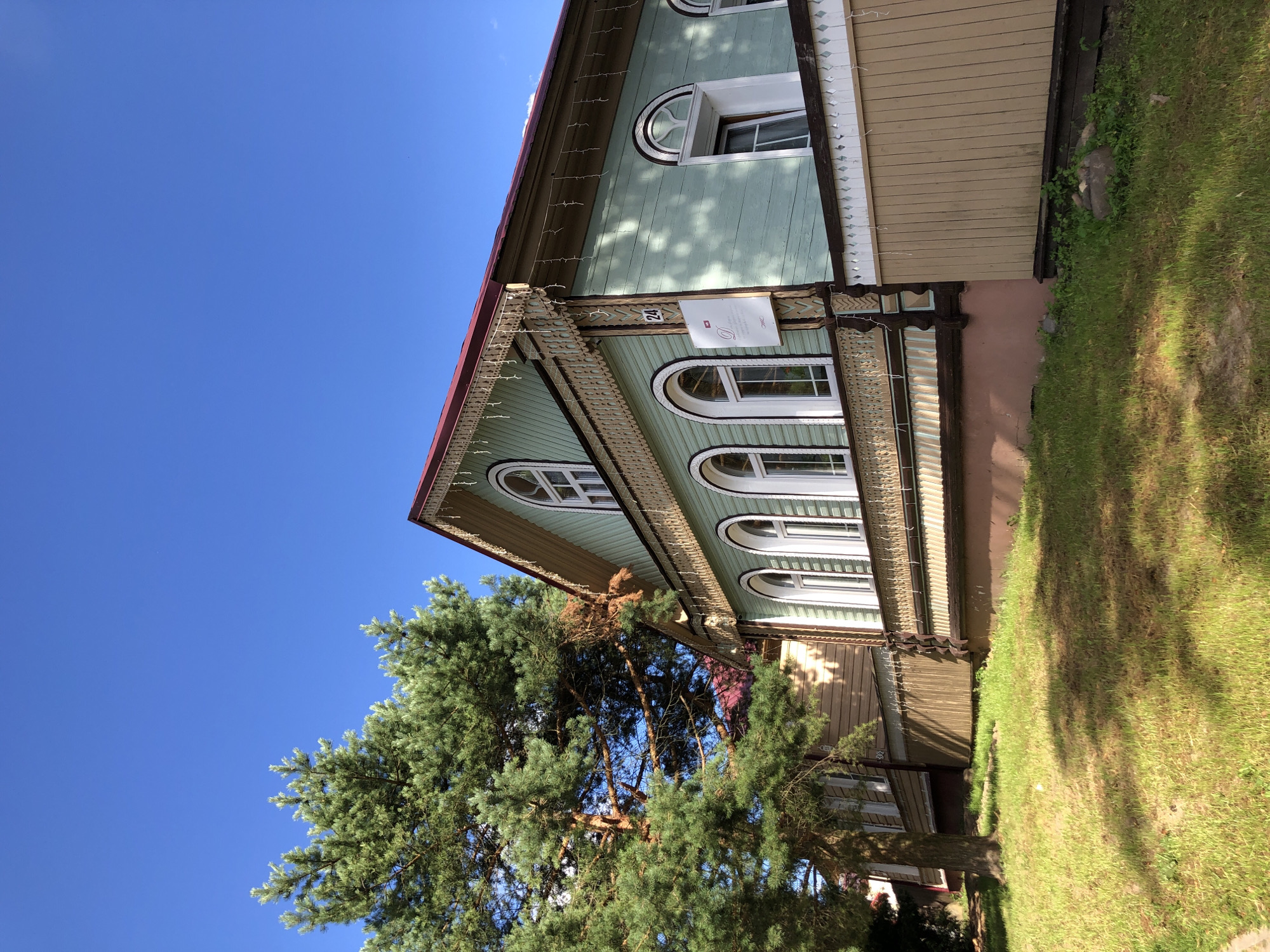 In Myshkin, Russia, a traditional wooden house painted light teal stands under a clear blue sky. The house features ornate wooden trim along the roofline and around its arched white-framed windows. A large evergreen tree with lush green needles is situated to the left of the house, casting dappled shadows on the grassy lawn in front. The foundation of the house is a light brown color. A tan wooden fence runs along the front of the property. A street number, "24", is visible on a white sign attached to the side of the house, next to another sign with text and a logo. The scene is bathed in bright sunlight, indicating a warm, clear day.