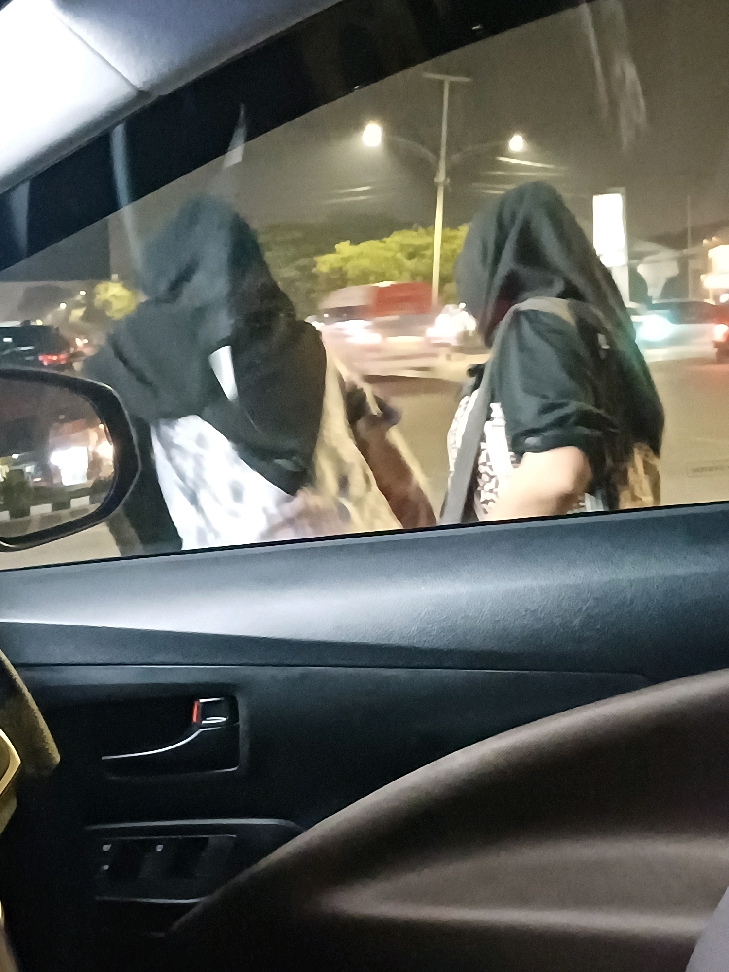The image is an indoor shot, taken from inside a vehicle looking out the window at night. Two individuals wearing black hooded garments and headscarves are visible outside the car. One individual is positioned to the left, with their back to the camera, and appears to be holding or wearing a white garment with a patterned design. The other individual is to the right, also facing away, and wearing a black jacket over a patterned garment. Streetlights illuminate the scene, casting a glow on the wet pavement, suggesting it may have recently rained. Cars and a lamppost are visible in the background, indicating an urban environment. The interior of the car is visible in the foreground, showing the door panel with a handle and what appears to be a window control. The overall impression is a candid snapshot of two people by the roadside at night, likely in Dumai City, Indonesia, given the context.