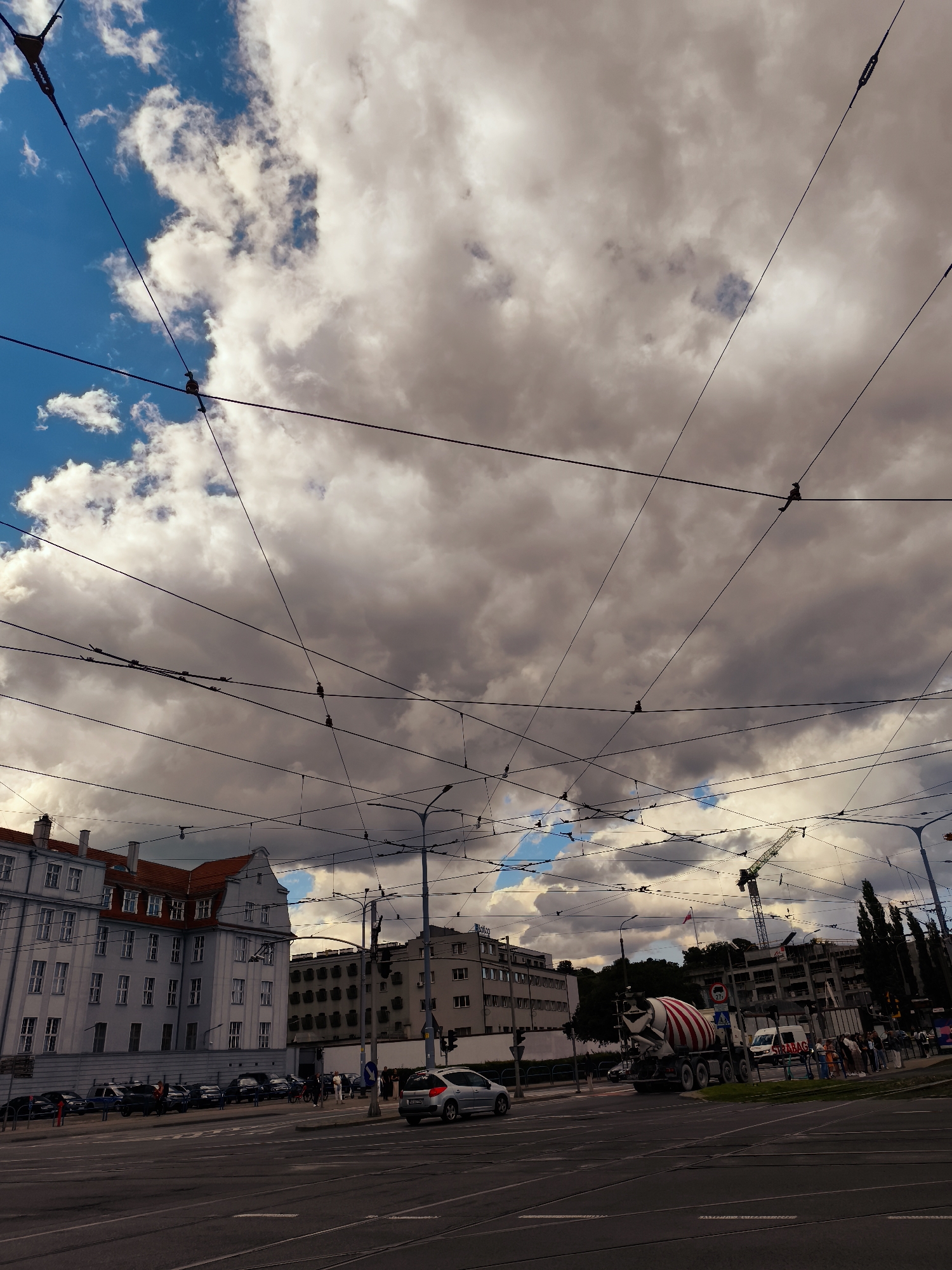 This image depicts an overcast sky over a city intersection in Gdańsk, Poland. The sky is a dramatic mix of dark, heavy clouds and patches of bright blue, suggesting a dynamic weather front. A complex network of overhead wires, likely for public transportation such as trams, crisscrosses the frame, dominating the upper portion of the image.
In the foreground, a street intersection is visible, with road markings and what appear to be tram tracks. On the left, a multi-story building with a red-tiled roof and numerous windows stands prominently. To its right, other buildings of varying architectural styles and heights are visible, some appearing to be under construction or renovation, indicated by a prominent green construction crane in the mid-ground.
Several vehicles are present on the road. A silver car is driving away from the viewer on the right side of the intersection. A large concrete mixer truck with red and white stripes is turning at the intersection. A white van and various other cars are parked or driving on the street. A small group of pedestrians is visible on the sidewalk near the construction site. The overall impression is one of an active urban environment under a temperamental sky.
