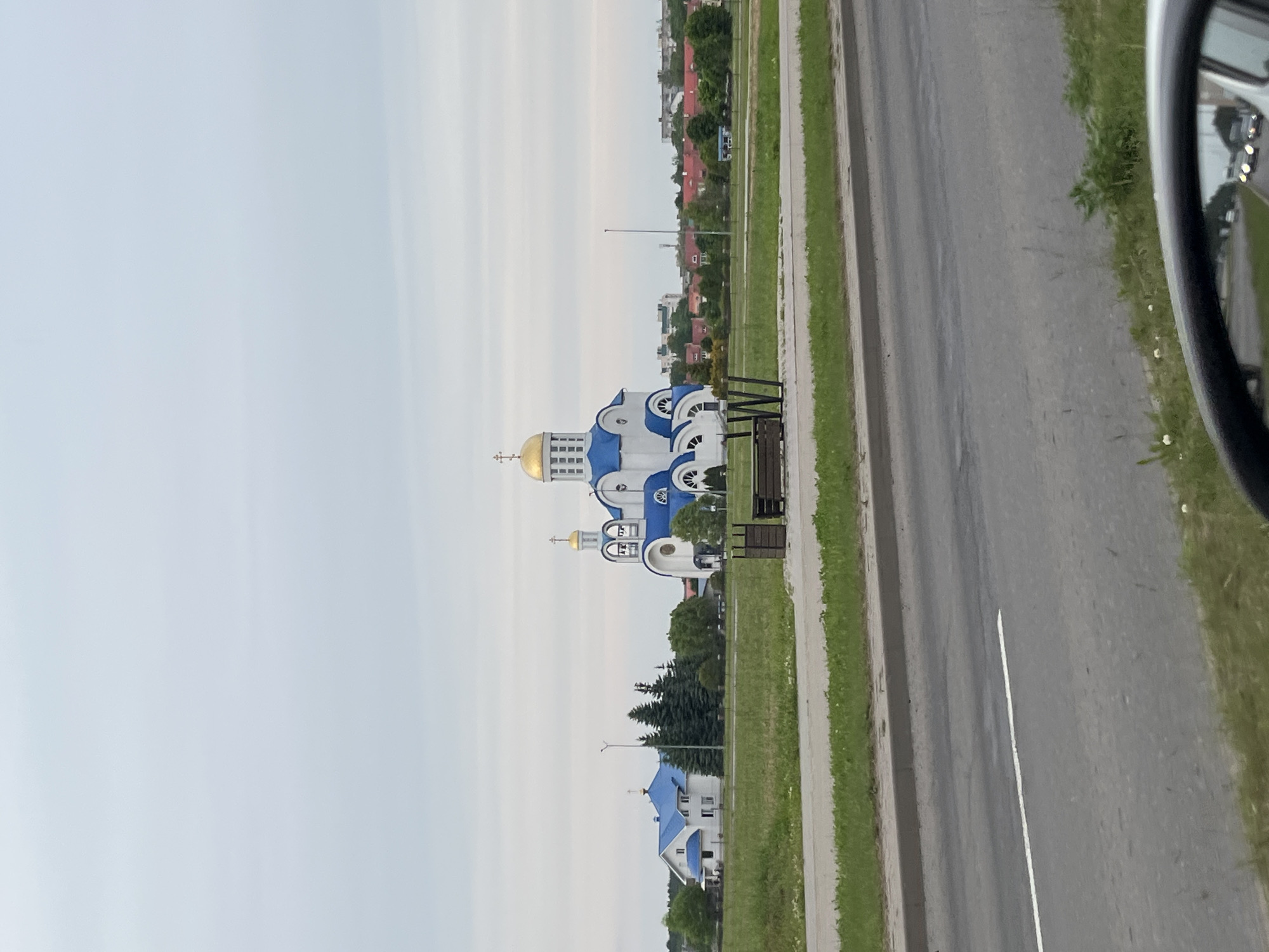 The image displays the Orthodox Church of the Intercession of the Holy Virgin in Zhodzina, Belarus. The church, positioned centrally, is a prominent structure featuring white walls, blue conical roofs, and multiple golden onion domes topped with Orthodox crosses. A larger golden dome caps the main central section, with smaller golden domes on adjacent towers. To the left of the church, a white two-story building with a matching blue roof stands adjacent to a cluster of dark green coniferous trees. The immediate area surrounding these structures is a vast, flat expanse of green grass. In the middle distance, behind the church, a townscape with numerous residential buildings characterized by red roofs is visible. The sky is uniformly light grey and overcast, with no discernible clouds.
In the foreground, a paved asphalt road extends horizontally across the lower third of the image, marked by a single white solid line. A grassy verge separates the road from the field where the church is located. On this grassy verge, slightly to the right of the image center, a dark wooden park bench and a matching waste bin are present. The lower right corner of the image captures a portion of a car's side-view mirror, reflecting blurred headlights of vehicles traveling behind, indicating the photograph was taken from inside a moving or stationary automobile. No people are visible in the scene.