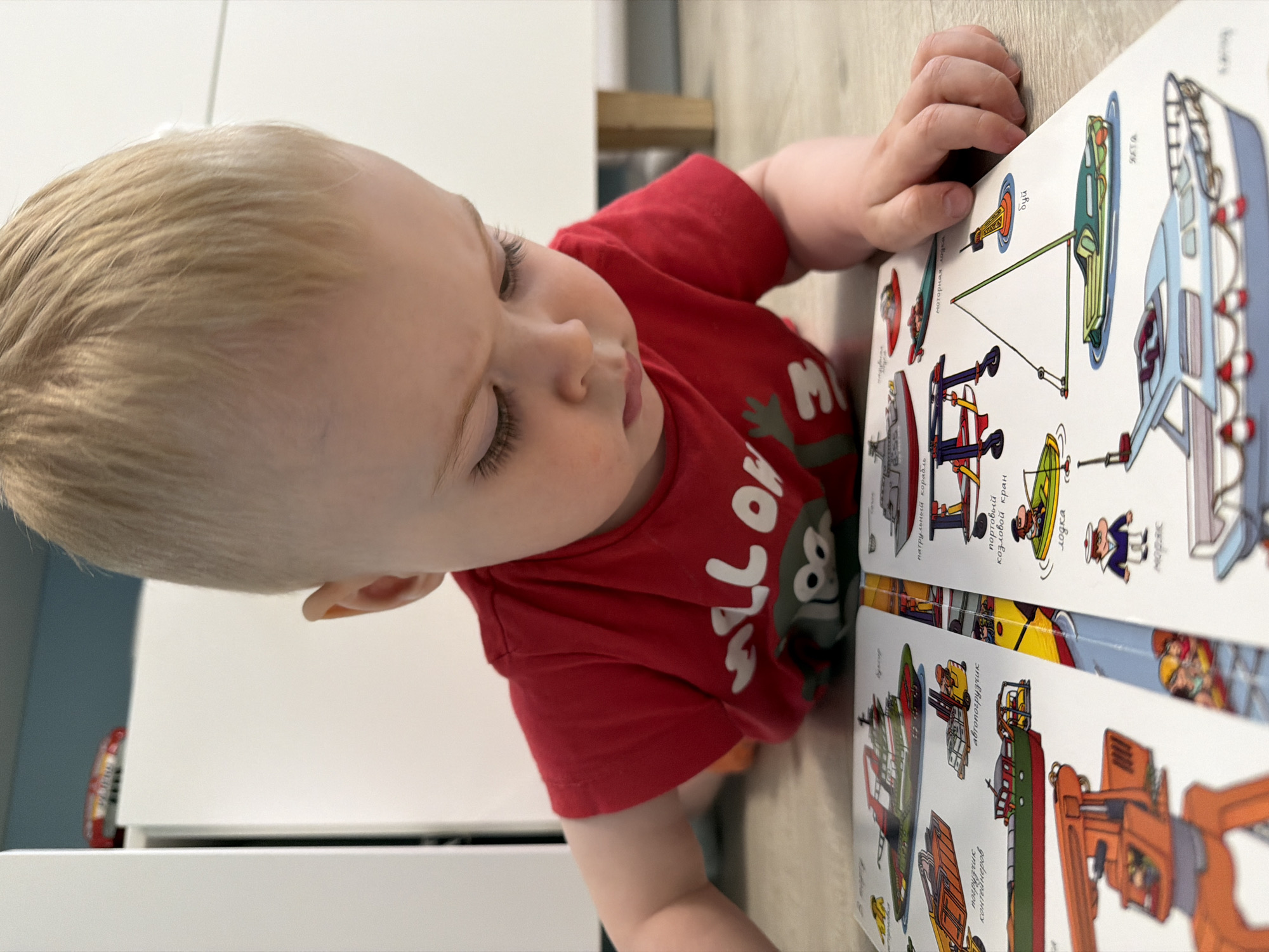 A young child with blond hair, wearing a red t-shirt, is lying down and looking at a book. The book features colorful drawings of boats and vehicles, along with Russian text labels such as "погрузчик контейнеров," "автопогрузчик," "патрульный корабль," "портовый козловой кран," "лодка," "моторная лодка," "буй," and "SATA." The child's hand is resting on the edge of the book. The scene appears to be indoors, with a white cabinet visible in the background.
The location of Votkinsk, Russia is not discernible from the image.