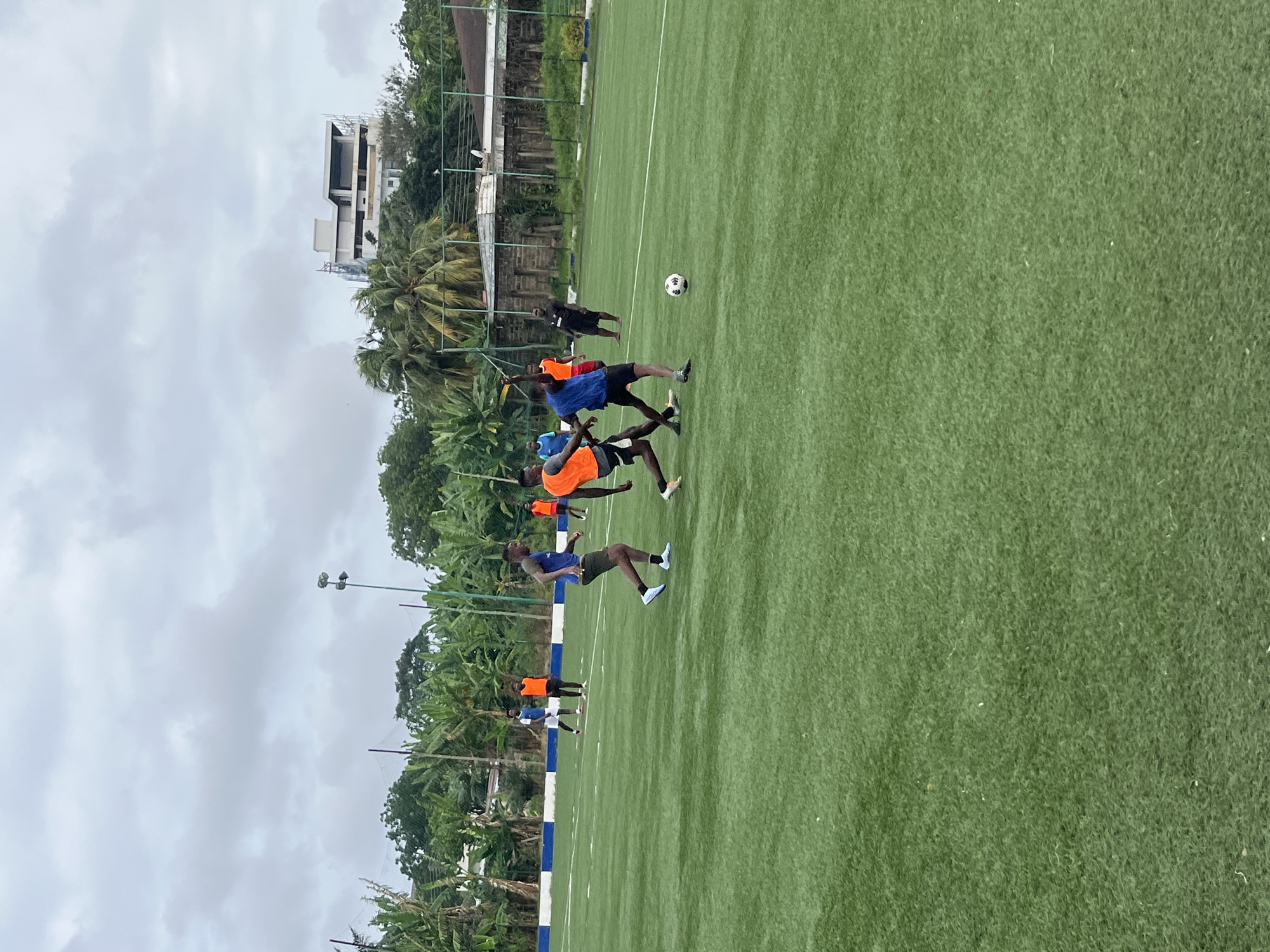 A group of young men are playing soccer on a green turf field. The field appears to be a sports facility with white lines marking boundaries and a blue and white border. In the background, there are trees, including banana trees, and a building under construction. The sky is overcast. The scene is likely in Accra, Ghana, given the presence of tropical vegetation and architectural styles.