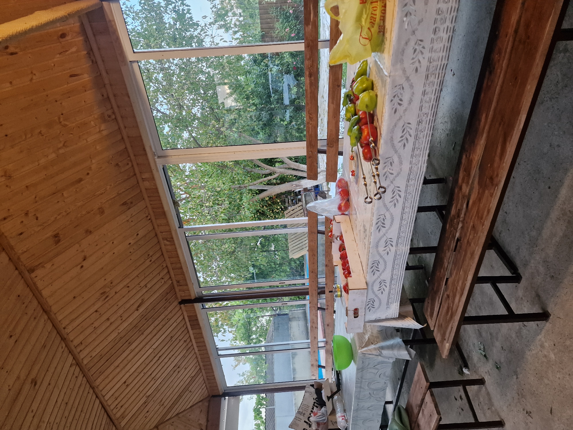 The image depicts an outdoor covered patio, possibly in Kursavka, Russia, with a wooden paneled ceiling and large glass windows overlooking a backyard. Several tables are set up, covered with tablecloths, and laden with food and preparation items for a meal. In the foreground, a wooden bench is visible. On one table, there are skewers with tomatoes and green peppers, suggesting preparations for grilling. Another table holds a box of tomatoes and a green bowl. The backyard visible through the windows contains trees, fencing, and what appears to be a swimming pool. The overall scene suggests a casual outdoor gathering or meal preparation.