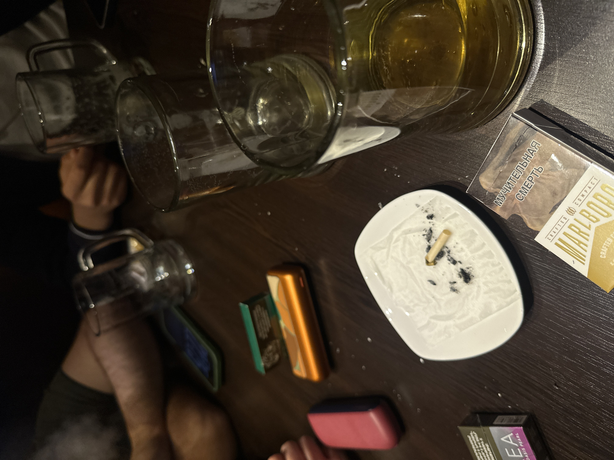 The image depicts a table scene, likely inside a bar or pub based on the presence of beer mugs. There are three beer mugs on the table, one full, one half-full, and one empty. An ashtray with cigarette butts and ash sits on the table next to a pack of Marlboro cigarettes with Russian text. Other items on the table include what appears to be a vape device, another pack of cigarettes (possibly of a different brand), and a pink object. Two individuals are partially visible, one holding a beer mug and the other with their hand on the table. The location and city cannot be determined from the image.