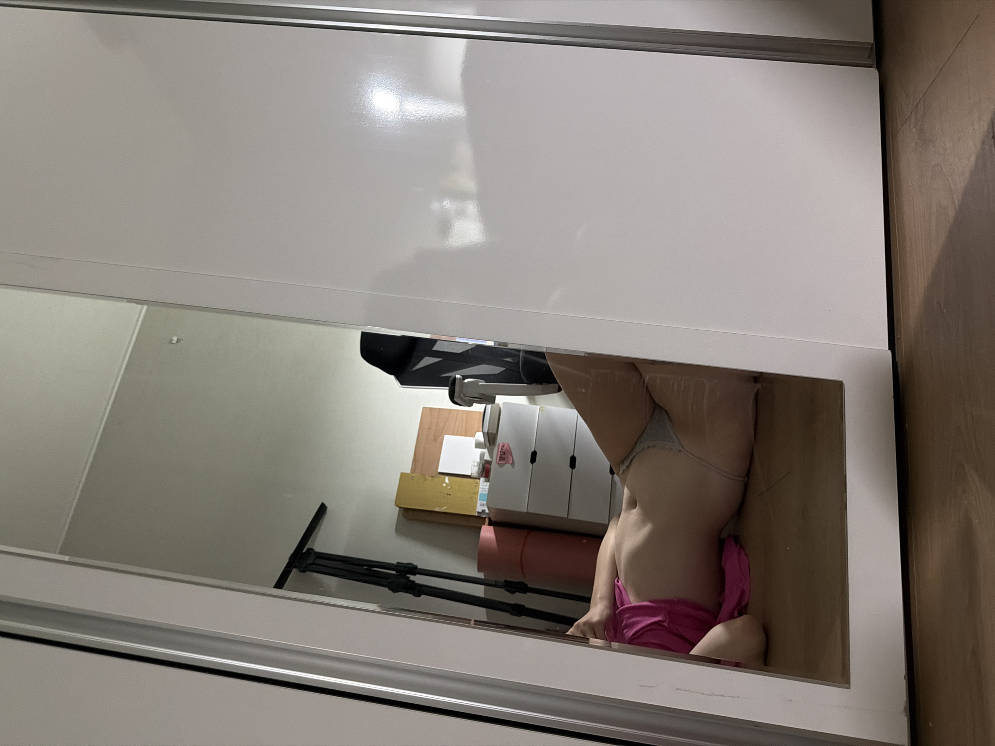 The image shows a person lying on the floor, partially visible in a mirror reflection. The person is wearing a pink top and grey underwear. The environment appears to be a room with a closet and some storage items. It is not possible to determine the exact location in Dongducheon-si, South Korea, or provide any further details about the scene.