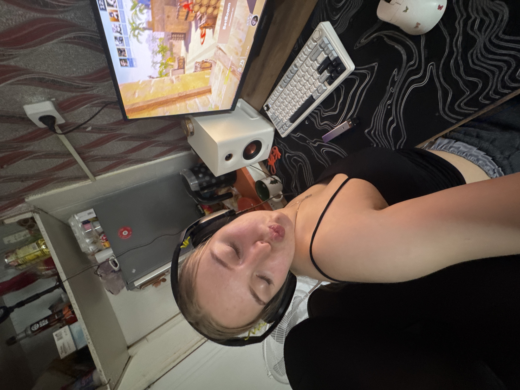 A person wearing headphones is sitting in a black chair in a room with a monitor displaying a video game. A white keyboard and a computer mouse are on a desk with a black mousepad. A white speaker is next to the monitor. The room has a shelf with bottles and containers. An electrical outlet is visible on the wall. The location appears to be a residential setting and the geographical location cannot be determined from the image.
