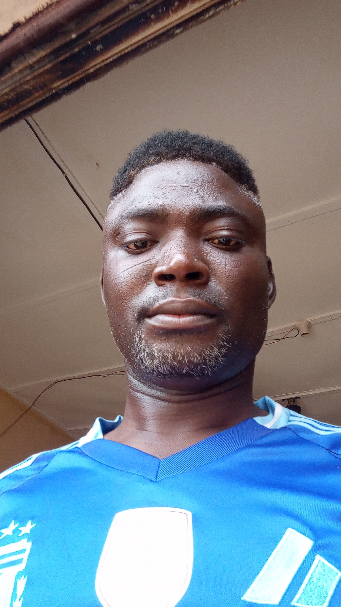 A close-up, low-angle selfie of a Black man with dark skin and a partially graying beard. He has short, dark hair and a small scar near his left eye. He is wearing a blue sports jersey with white accents. The background shows a ceiling with some wiring and wooden trim. There is no explicit indication the location is Sabon Tasha, Nigeria.