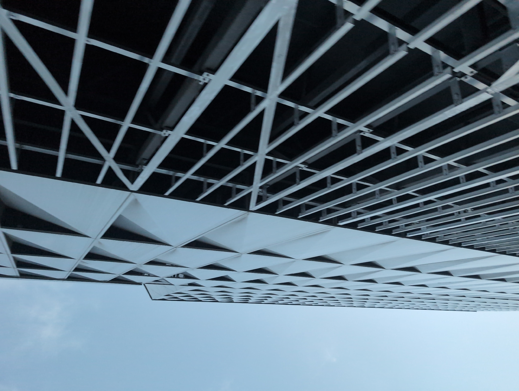 This is a low-angle, exterior, daylight shot of the modern, geometric facade of a building, likely a parking garage, with a clear blue sky in the background. The building's exterior is composed of two distinct sections. On the left, a large section features a repeating pattern of diamond-shaped indentations or panels, creating a textured, three-dimensional surface. This section is illuminated by natural light, revealing its smooth, light-colored material. To the right of this section, a second part of the facade is visible, characterized by a grid of thick, white metal bars forming diagonal and horizontal lines against a dark background. This appears to be an open-air structure, possibly a parking level with railings or protective barriers. The perspective emphasizes the upward lines and angles of the architecture, with the building appearing to recede into the sky. No people are visible in the image. The environment is an urban setting, under a bright, cloudless sky. The architecture suggests a contemporary design, possibly located in a city like Kyiv, Ukraine, known for its modern architectural developments.