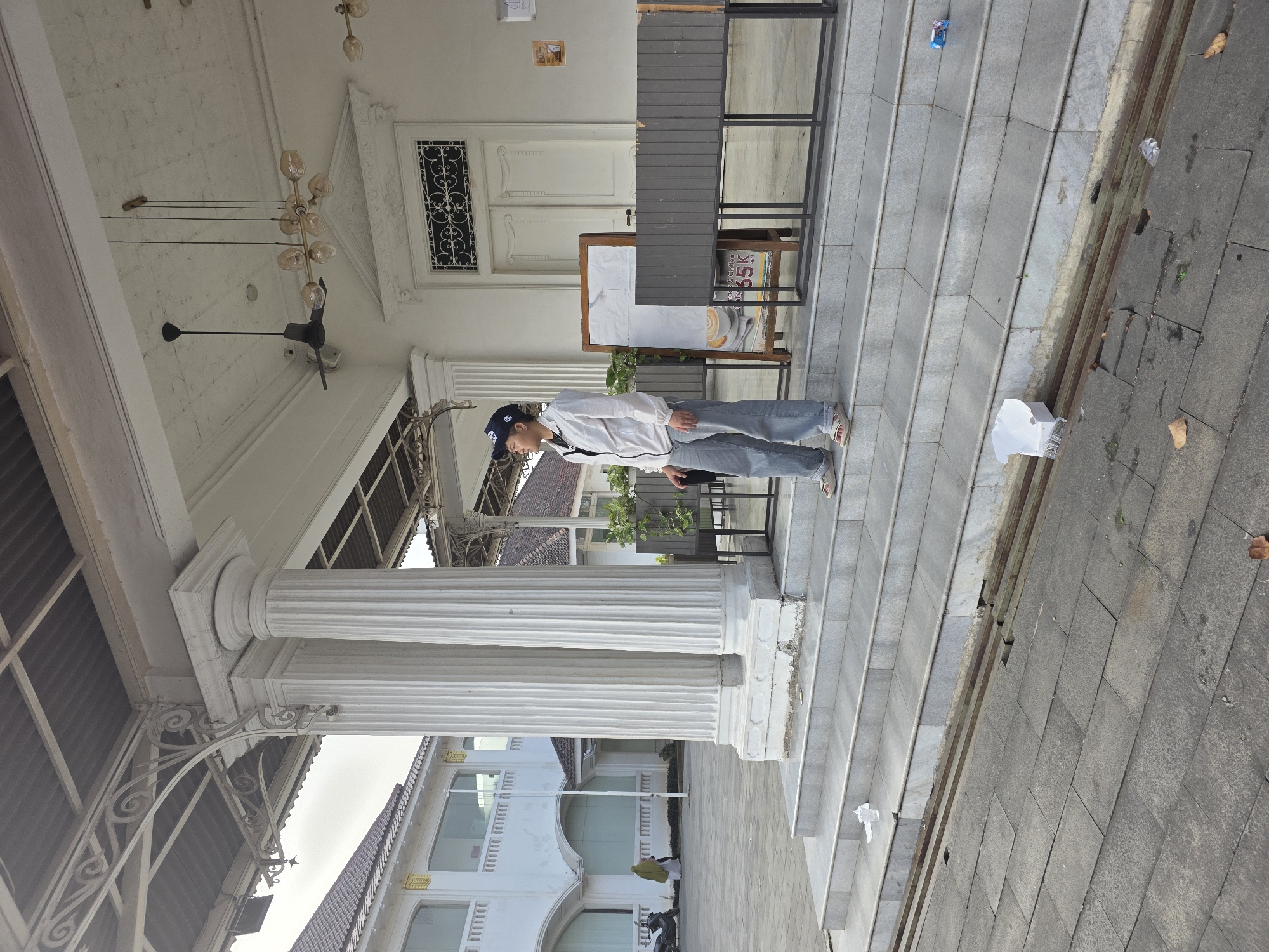 A person stands on a set of gray stone steps outside a white building. The building has large, fluted white columns supporting a covered walkway. The person, wearing a white jacket, blue jeans, a blue baseball cap, and light-colored sandals, is looking to the right. To the right of the person, there is a wooden easel with a white paper taped to it, and a dark gray, slatted structure. A door with ornate white trim is visible behind the easel. A ceiling fan and a modern chandelier hang from the ceiling of the walkway. In the background to the left, another white building with arched windows and a tiled roof is visible, and a person wearing a green headscarf and a scooter are present. The ground in front of the steps is paved with gray stone tiles. Several pieces of litter, including a white takeout box, are scattered on the steps and the pavement. The overall scene suggests an outdoor public space, possibly a cafe or a historic building in Tambun, Indonesia.