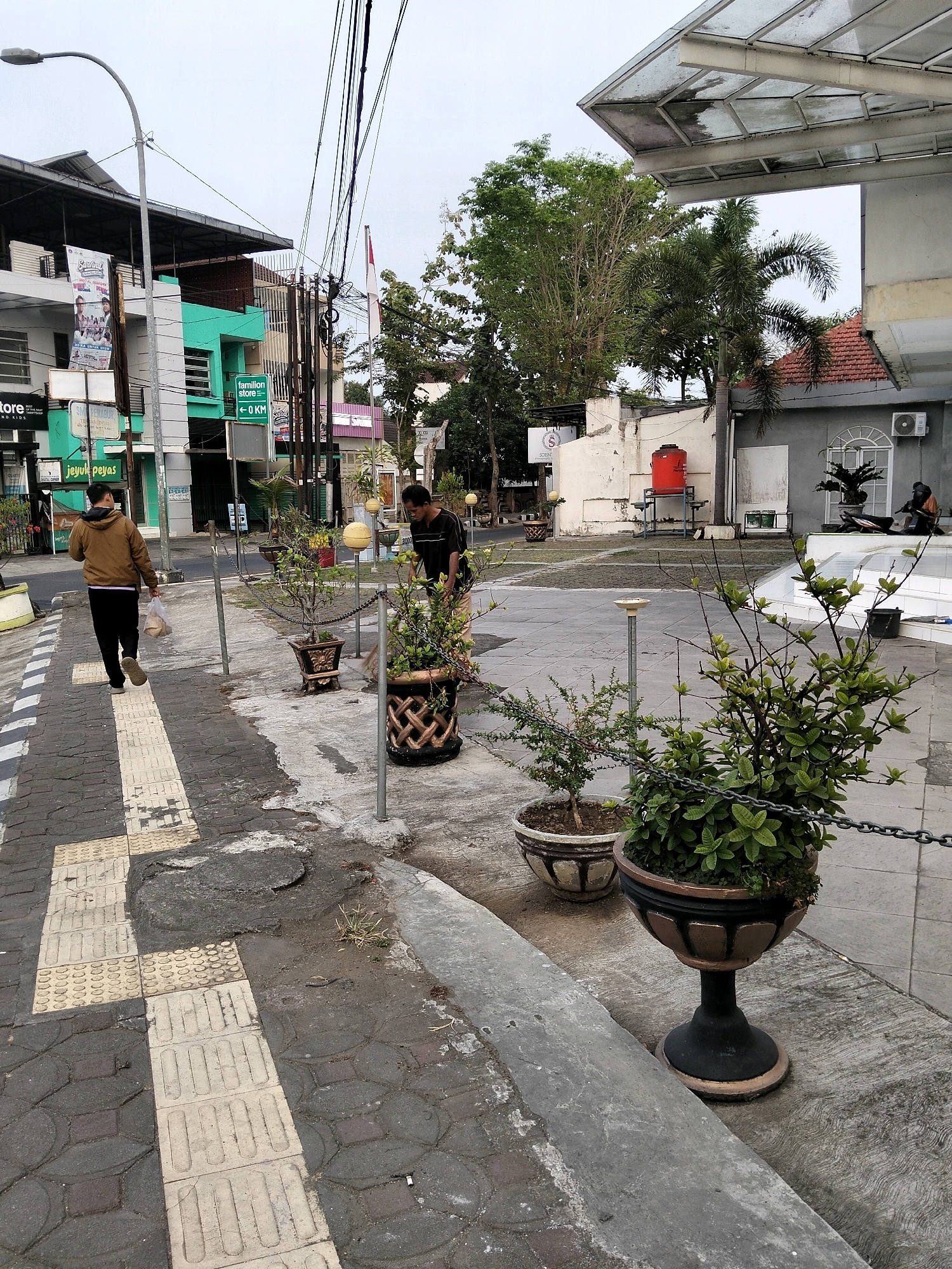 The image depicts a street scene in Purworejo, Indonesia, likely near a commercial area. In the foreground, a person walks away from the camera on a sidewalk paved with textured blocks for tactile guidance. To the right, two men are tending to plants in decorative pots. One man is sweeping, while the other appears to be working with a plant. In the background, buildings with storefronts and various signage are visible, along with trees and utility poles. A red water tank is situated near a building. The overall atmosphere suggests an ordinary day in a city.