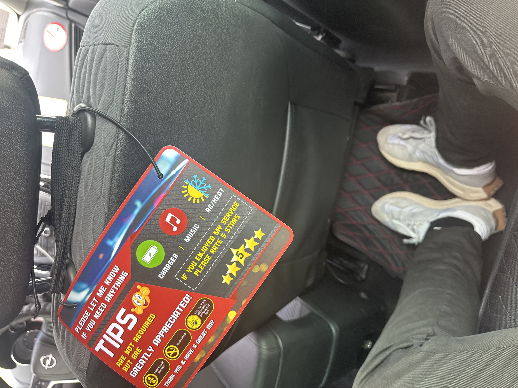 A photograph taken inside a vehicle features a sign hanging from the back of the driver's seat which reads, "PLEASE LET ME KNOW IF YOU NEED ANYTHING" and "TIPS ARE NOT REQUIRED BUT ARE GREATLY APPRECIATED!" It also indicates the availability of a charger, music, and AC/heat. The sign further encourages the passenger to rate the service 5 stars if they enjoyed their ride. Adjacent to the seat, a person is visible wearing grey sneakers, black pants, and black socks. A diamond pattern is visible on the car's floormat.