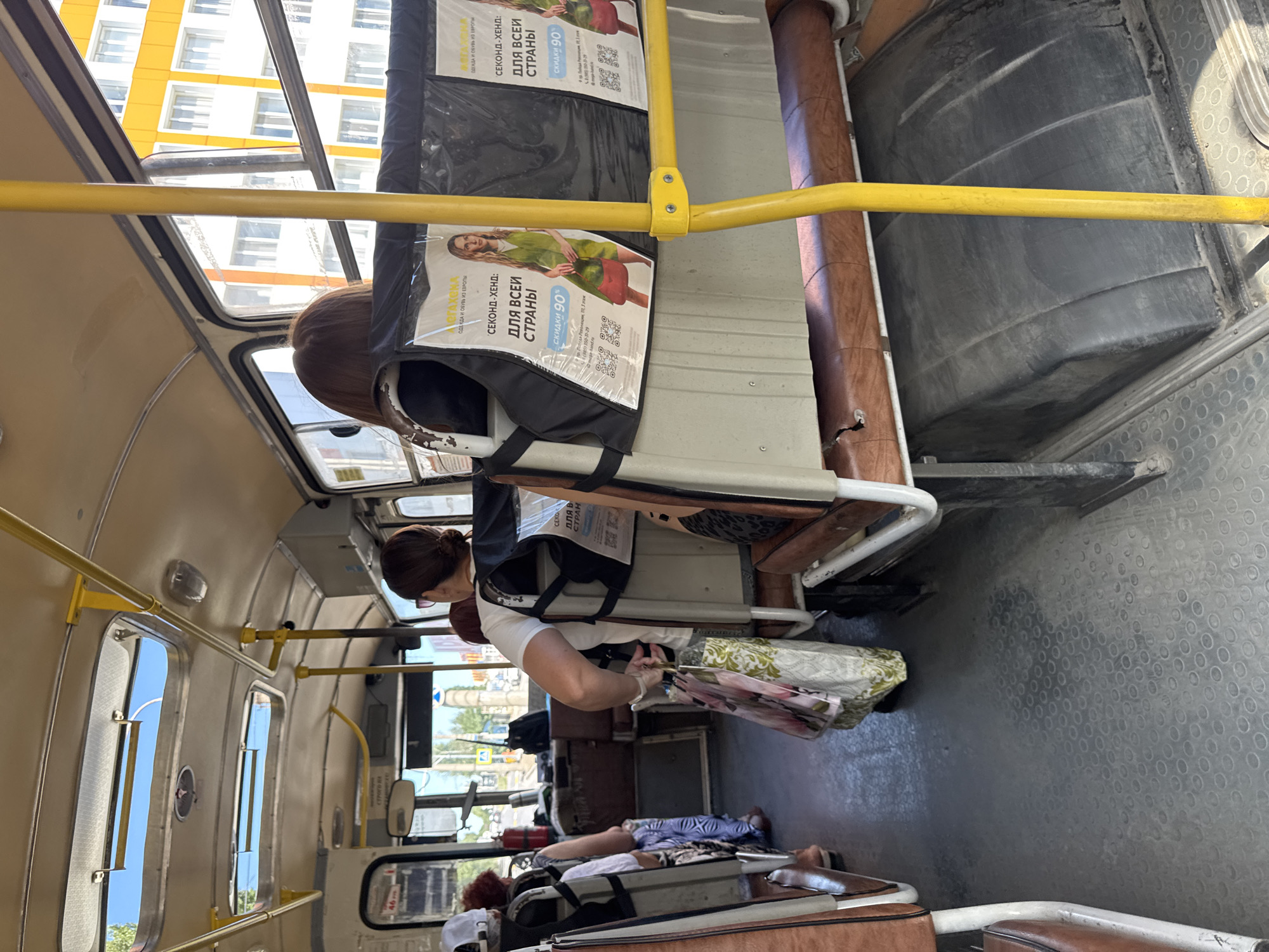 An interior view of a bus in Shakhty, Russia, shows passengers seated and standing. The bus features rows of brown vinyl seats and yellow support poles. Overhead, there are skylights revealing a blue sky. Advertisements for a second-hand clothing store are displayed on the seatbacks, featuring images of women in clothing and offering a 90% discount. The floor of the bus is gray and textured. In the foreground, a woman is holding shopping bags. Outside the bus windows, buildings and trees are visible.