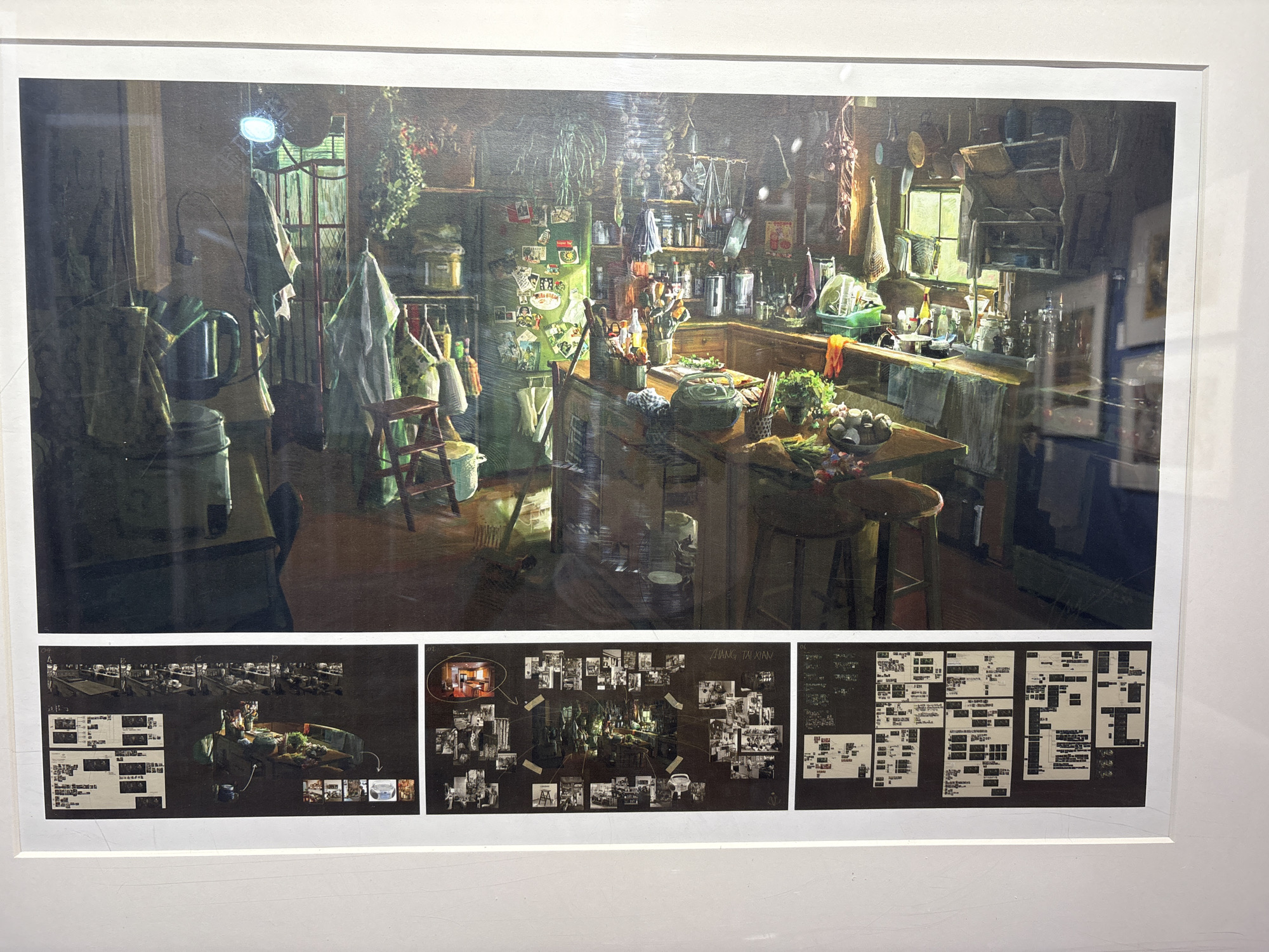 The image shows a framed artwork. The artwork consists of a large, painterly image of a cluttered kitchen. Above the kitchen's working spaces and sink is shelving lined with jars, containers, and dried herbs. Hanging from the ceiling are garlic braids and other provisions. Light streams in from a window, illuminating a stool and vegetables on the countertop. Below the main image are three smaller, dark-backgrounded compositions. The leftmost composition includes a time-lapse sequence above a grid of images with accompanying text. The central composition features a diagram linking a central kitchen image to related images placed around it. The rightmost composition shows a dense grid of text and image snippets. The artwork is displayed in a white frame and hung on a white wall. The photo was probably taken in a museum or gallery; however, there is nothing to suggest this is located in the city of Nan'an District, China.