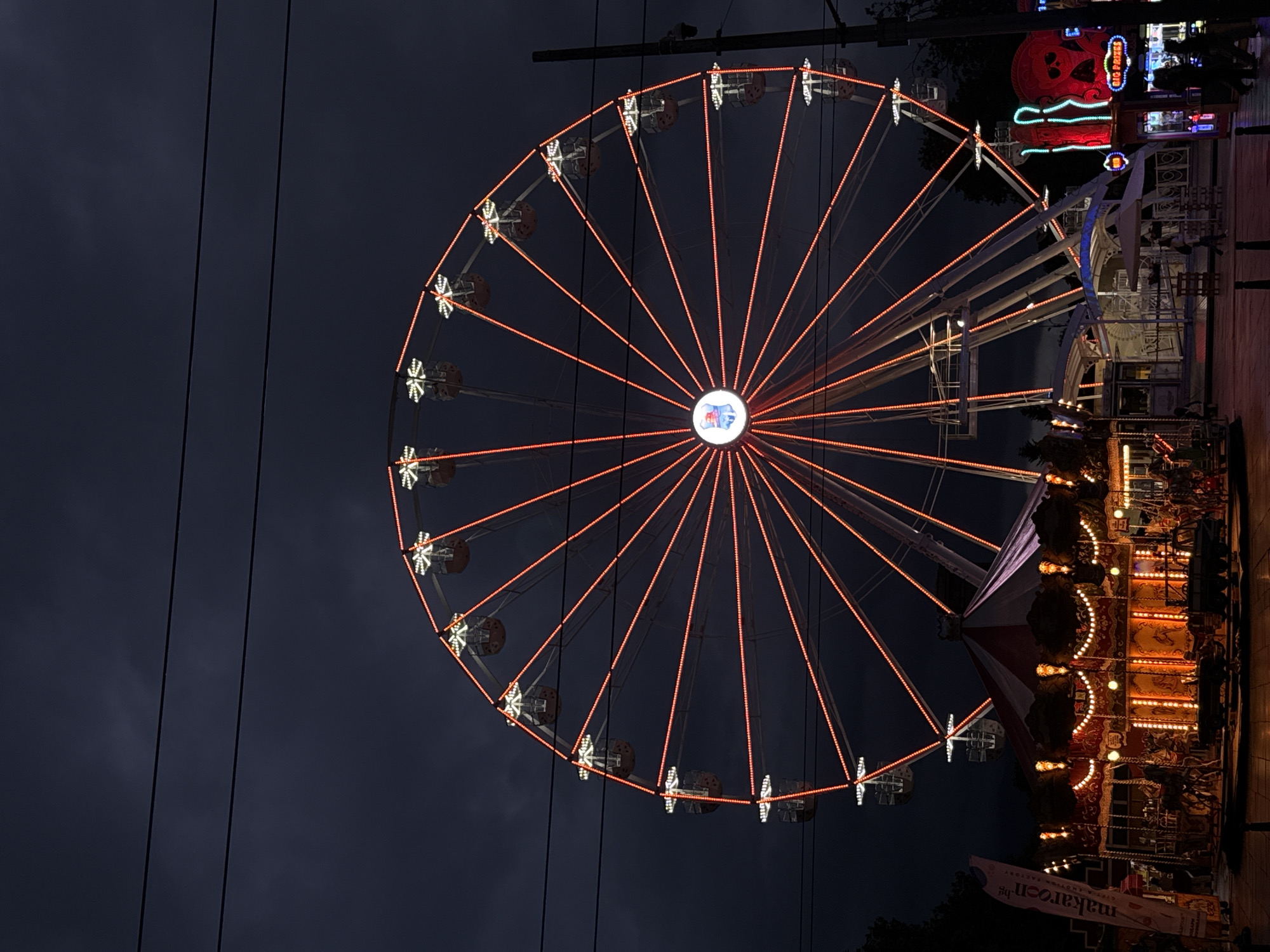The image shows a Ferris wheel illuminated with orange lights at night. The wheel has a central logo and white lights around its perimeter. The background is a dark sky. To the right, there are other illuminated structures, including a carousel and a building with decorative red and green lights. A sign with the text "makarell.bg" is visible at the bottom right. The image is likely taken in Sofia, Bulgaria, based on the location mentioned in the query.