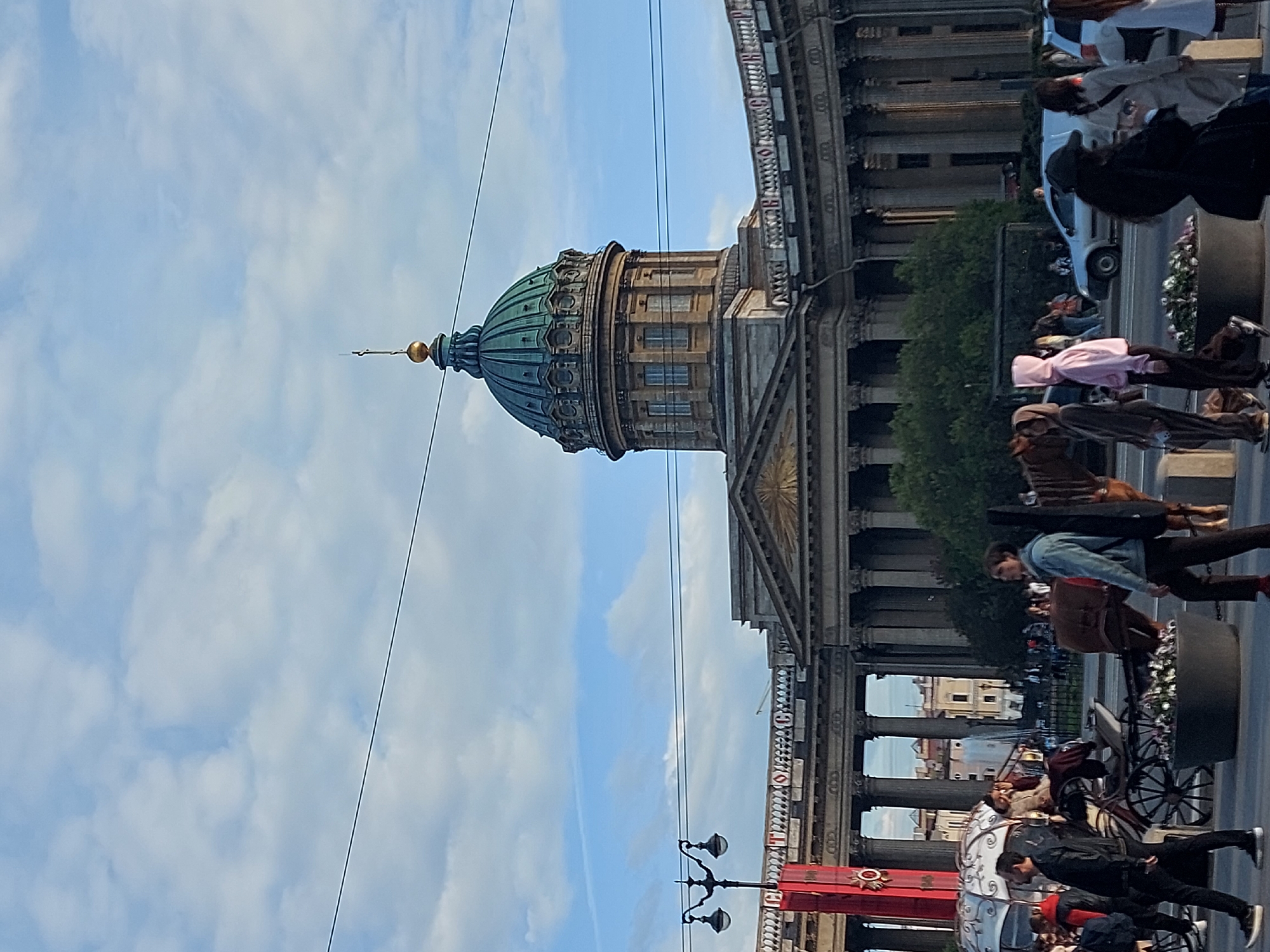 The image captures the Kazan Cathedral in Saint Petersburg, Russia. The cathedral's distinctive green dome and neoclassical façade stand out against a partly cloudy blue sky. The cathedral's architecture features a colonnade extending along the sidewalk, and the foreground is bustling with people. Some are walking and others are engaging in tourist activities, such as riding in horse-drawn carriages. Street lamps are visible.