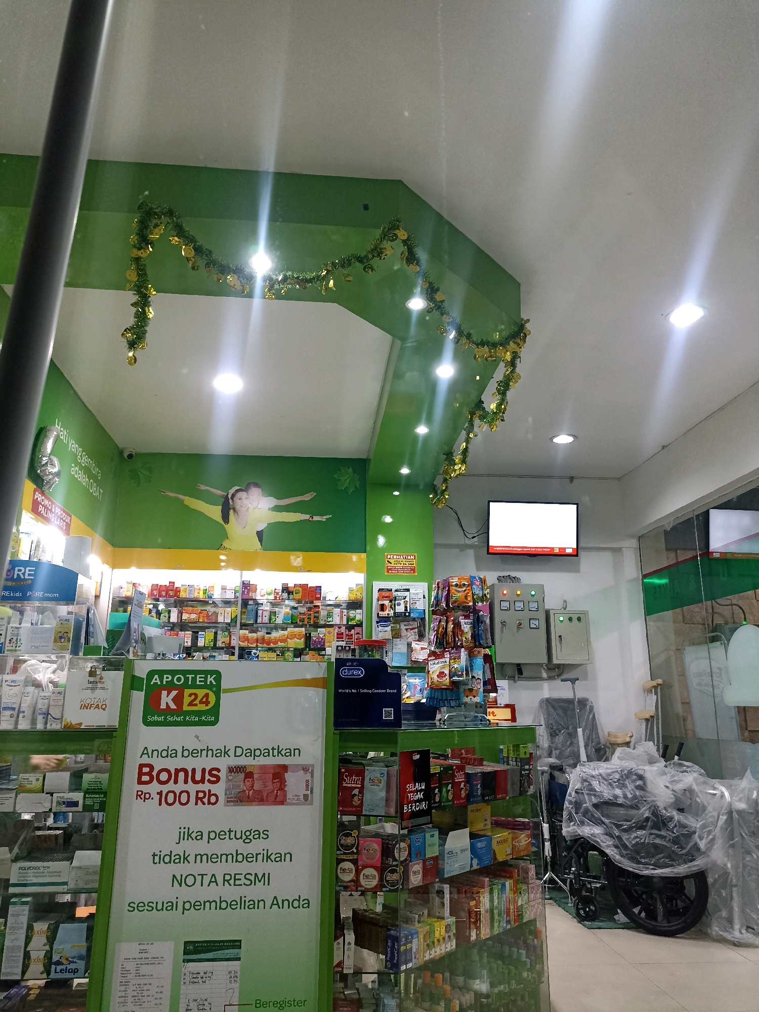 The image depicts the interior of an Apotek K24 pharmacy, likely located in Lamongan, Indonesia, given the signage. The pharmacy features a green and white color scheme, with shelves stocked with various pharmaceutical products. A large banner prominently displays "APOTEK K24" and a promotional offer for a bonus of Rp. 100,000 if a formal receipt is not provided.
In the background, a large mural of two people with outstretched arms is visible on the green wall. Above, festive green garlands with golden bells are strung across the ceiling. A television screen is mounted on the wall, displaying a blank white screen with some text at the bottom. Electrical control panels are also visible.
In the foreground, a glass display case showcases more products. To the right, a wheelchair is covered with plastic sheeting, along with crutches, suggesting the availability of medical equipment for sale or rent. The overall impression is of a well-stocked and organized pharmacy.