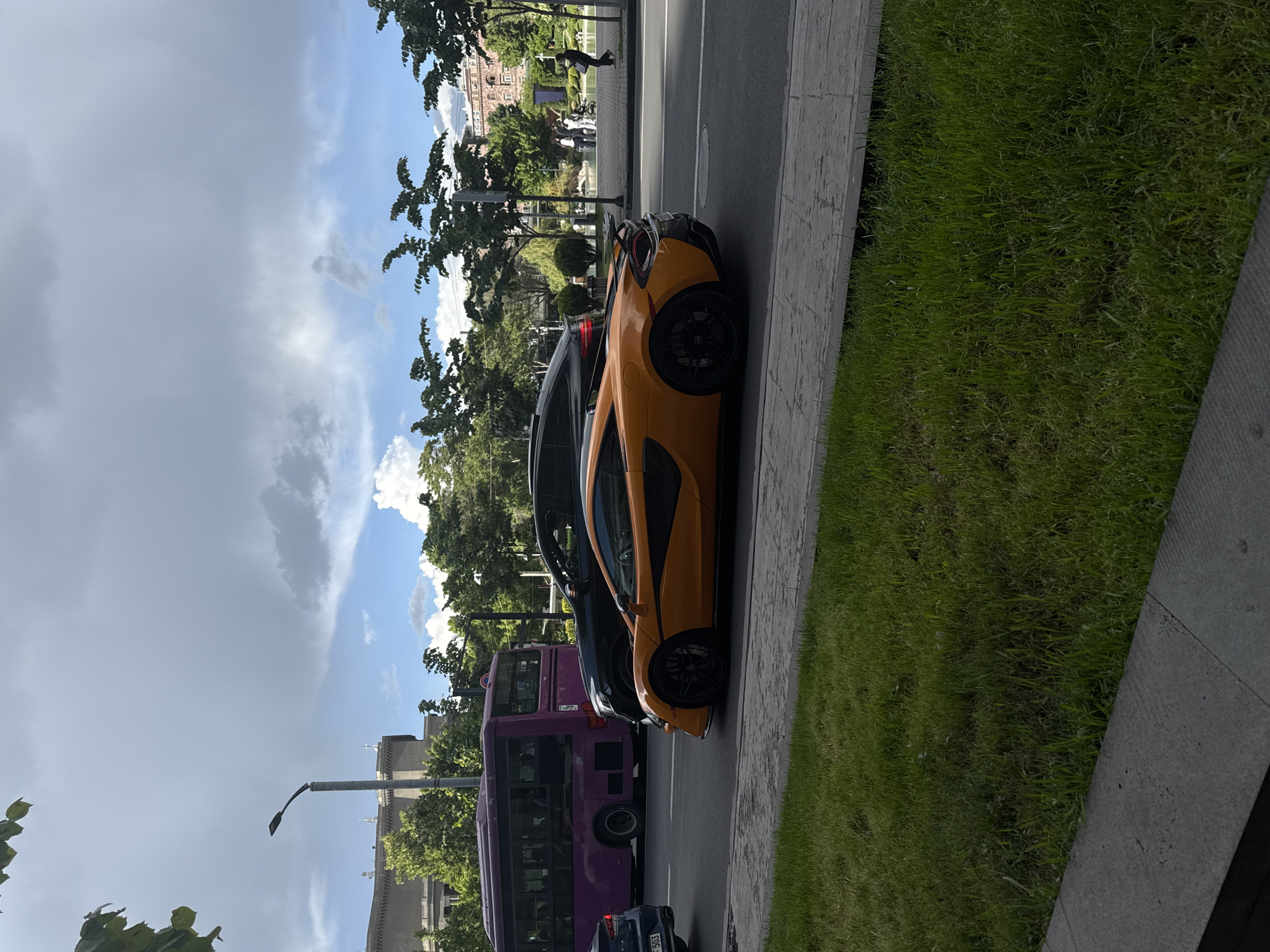 A medium eye-level shot depicts a street scene in Yerevan, Armenia. A vivid orange McLaren sports car is parked adjacent to a sidewalk with a patch of green grass and concrete curb on the right. A dark-colored sedan is parked behind it, followed by a purple bus. A person is walking on the sidewalk on the left, near trees and buildings. The sky is partly cloudy. A street lamp is visible in the distance. The angle of the shot makes the scene appear tilted.