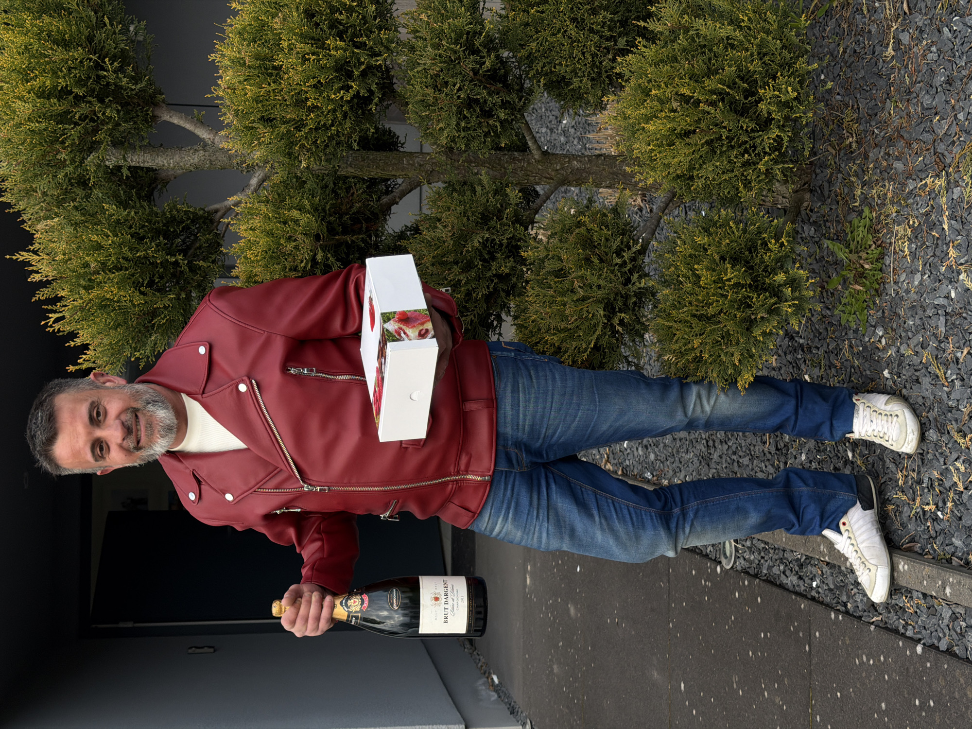 A male subject stands outdoors in Junglinster, Luxembourg, facing forward and smiling. He possesses a salt-and-pepper beard and short hair. He is wearing a red leather jacket with prominent zippers, a white crew-neck shirt, blue denim jeans, and white athletic shoes with dark soles. In his left hand (viewer's right), he holds a dark bottle labeled "Brut D'Argent Blanc de Blancs Chardonnay." In his right hand (viewer's left), he holds a white rectangular box, partially revealing multiple items with red toppings. The background consists of a dark gray building with an open door. To the subject's right, a trimmed coniferous shrub with green and yellow foliage grows from a bed of dark gray gravel. The subject stands on a dark paved surface with light specks, bordered by a concrete curb separating it from the gravel.