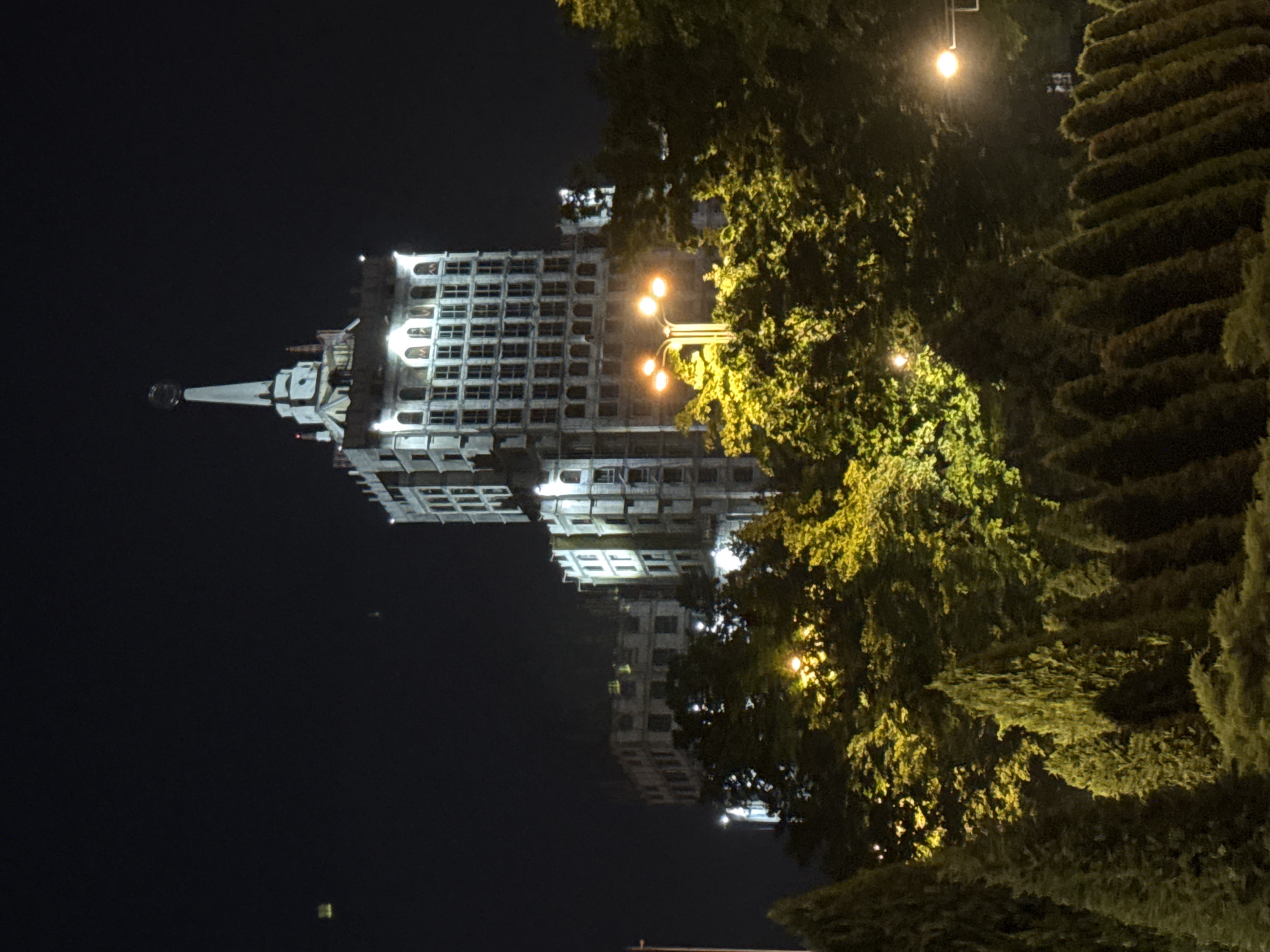 The image captures a striking urban scene at night in Bishkek City, Kyrgyzstan, centered on a tall, ornate building with a prominent spire. The building, rendered in a light, possibly white or grey material, is brilliantly illuminated against the dark sky, showcasing its multiple stories and numerous windows, some of which feature arched designs. Its imposing architecture suggests a significant civic or historical landmark, possibly from the Soviet era.
To the right of the building, a dense array of green foliage and trees partially frames the structure. These trees are vibrant, with patches illuminated by artificial light sources, casting a warm, yellowish glow that highlights their leaves and textures. A prominent streetlight with multiple lamps is visible in the middle-right, its bright light diffusing into the surrounding trees. Further to the right, neatly trimmed hedges or layered evergreen trees create a textured backdrop.
The time of day is clearly nighttime, characterized by the dark sky and the reliance on artificial lighting from streetlamps and the building itself. No people or discernible activities are visible in the tranquil scene. The image primarily focuses on the architectural presence of the illuminated building and the surrounding natural elements, creating a serene urban nightscape. There is no visible text.