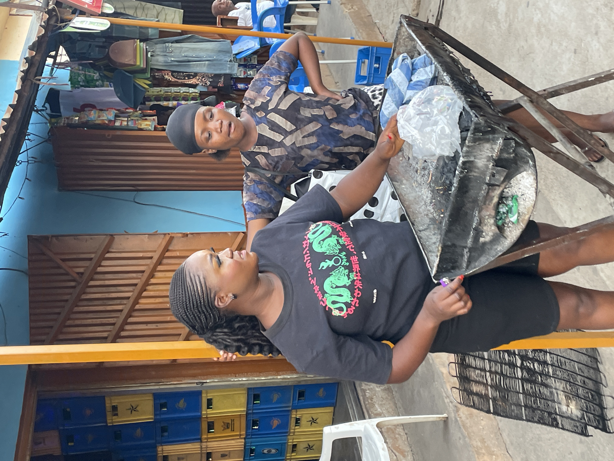 The image depicts a vibrant street scene in Jos, Nigeria, likely at a market or a street-side stall during clear daytime hours. The central focus is on two women engaged in an interaction, possibly a transaction or conversation, with a metal cart or tray between them.
In the immediate foreground, a woman stands on the left, looking towards the other woman with a faint smile. She wears a black t-shirt adorned with green dragon graphics and Japanese text. Her hair is styled in neat braids pulled back, and she is wearing a black skirt or shorts. Her left hand is near the metal cart, possibly holding a purple lighter or a small item.
To her right, a second woman stands, looking directly towards the first woman. She wears a patterned blue and brown long-sleeved top and a black headwrap. Her right arm is bent, with her hand resting on her hip, as she observes the interaction. The metal cart separating the two women appears utilitarian, containing items such as a white plastic bag and what might be charcoal or other goods, suggesting it's part of a vendor's setup.
In the background, behind the women, market elements are visible. Clothes hang on racks, indicating a clothing stall. Further back and to the lower left, stacks of blue and yellow plastic crates are piled, commonly used for beverages. A man is partially visible in the upper background, seated in a blue plastic chair, observing the scene. A light blue painted wall and a wooden slatted roof structure are also part of the background. The general atmosphere suggests a lively commercial area.
The visible Japanese text on the first woman's t-shirt reads:
- 「しません。ドラゴンは」 (Shimasen. Doragon wa) - "Don't. The dragon is"
- 「不安定で、世界は失われる」 (Fuan de, sekai wa ushinawareru) - "Unstable, the world is lost"
- 「終わる」 (Owaru) - "Ends"
These phrases likely complement the dragon imagery on her shirt.