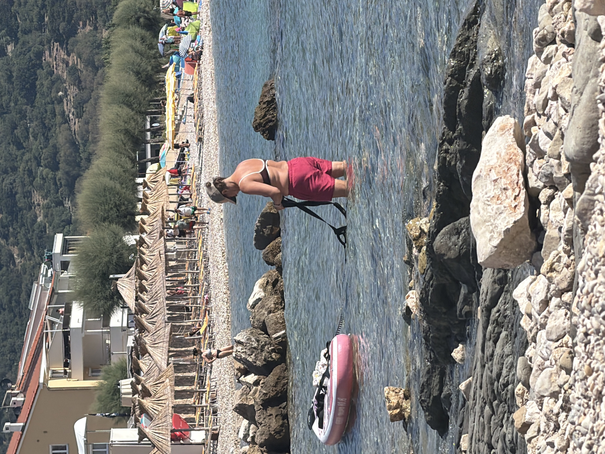 In the foreground of the image, a woman stands in shallow, clear blue water at a rocky beach in Općina Baška, Croatia. She is wearing a dark bikini top with a white strap and red shorts. She holds a black strap in her hands and appears to be adjusting or preparing it. To her left, a pink inflatable stand-up paddleboard with a black paddle and a white towel is tethered and floating in the water. The water is calm and slightly rippled.
The background reveals a bustling beach scene. Rows of thatched umbrellas are set up on a pebbled shore, with lounge chairs visible beneath them. Numerous people are scattered across the beach, some walking, others relaxing. Beyond the beach, a multi-story building with a red-tiled roof and balconies stands prominently. Further back, a dense line of green trees forms a backdrop against a hilly, vegetated landscape. The sky is clear and bright, indicating a sunny day.