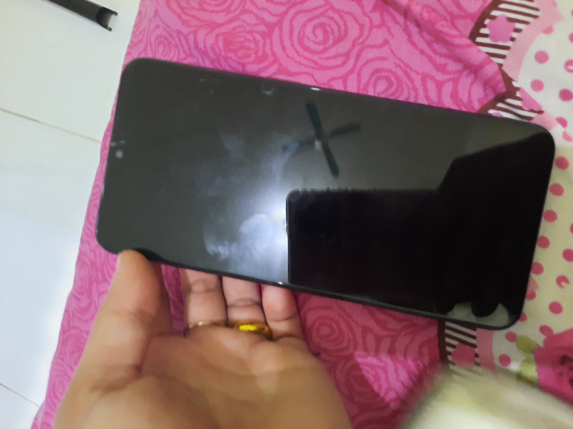 A person's hand, adorned with a gold ring on their ring finger, is holding a black smartphone. The phone is positioned horizontally, taking up the majority of the frame, with its screen reflecting a ceiling fan. The hand is resting on a pink pillow with a floral pattern. The background appears to be a bed or a soft surface covered with patterned fabric. The scene is indoors and has no discernible features that would place it in Lhokseumawe, Indonesia.