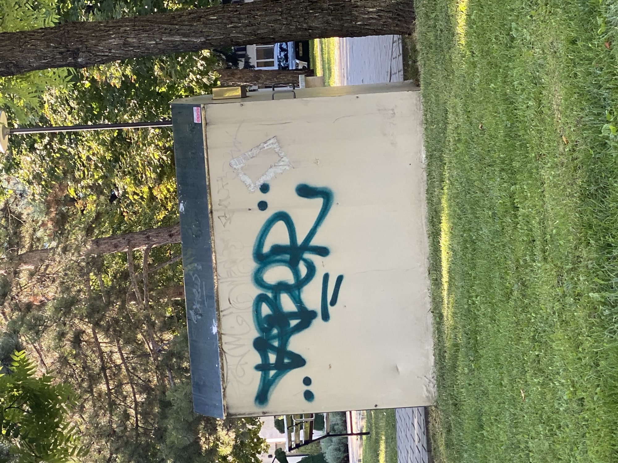 The image shows a light-colored, rectangular utility box located outdoors in a park-like setting. The box is covered with graffiti, the most prominent of which is a teal-colored tag written in a stylized font. The tag appears to say "ABOR". There are also other faint markings and a square piece of ripped paper attached to the box. The utility box is situated on a paved pathway, adjacent to a grassy area with trees in the background. A lamppost is visible behind the box, and part of a tree trunk is on the right side of the frame. In the background, a vehicle is parked on a street. The environment suggests a public outdoor space, likely in a city. Based on context, this scene is located in Chișinău, Moldova.