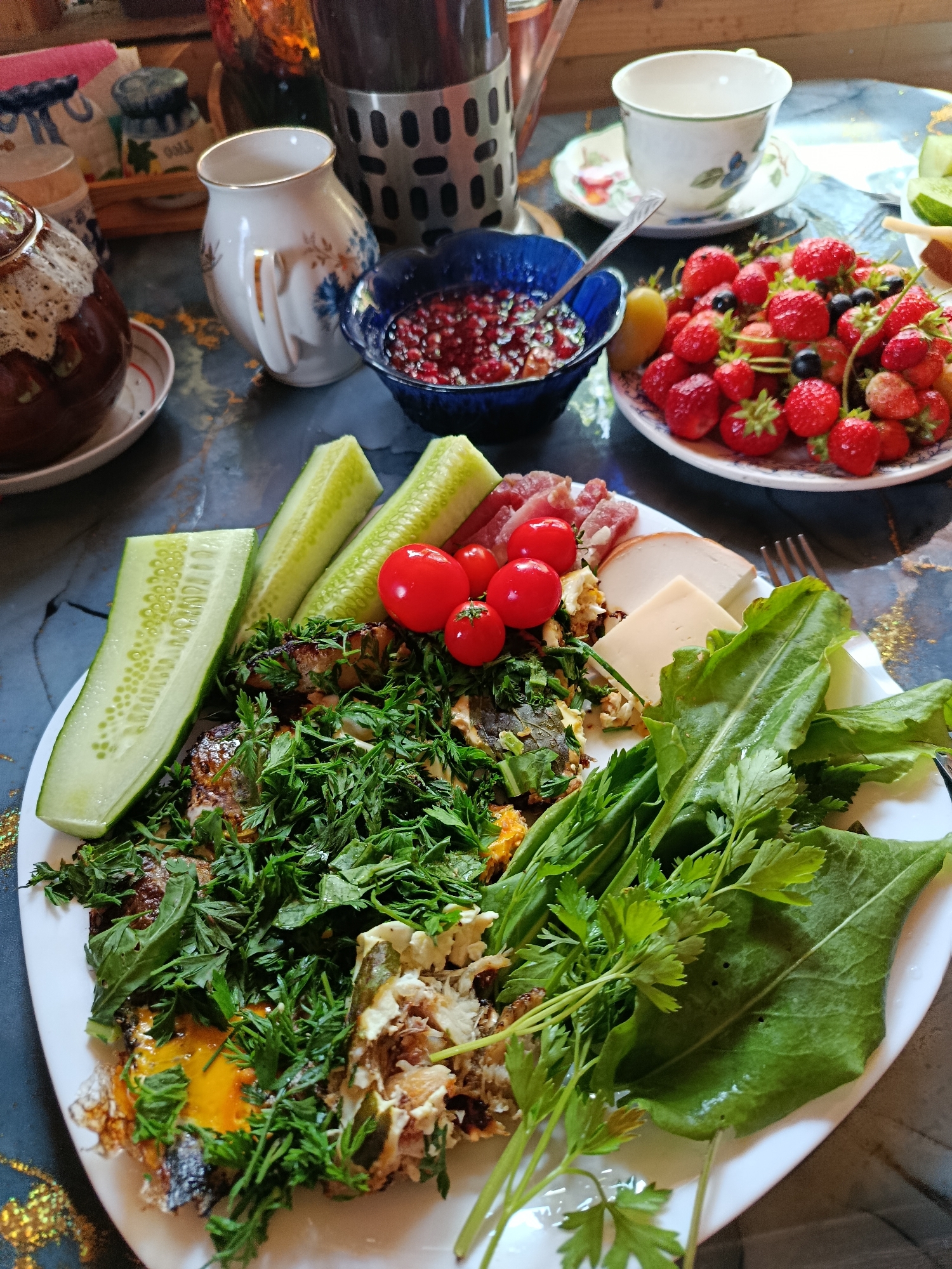 The image depicts a table setting with food and drinks, likely a breakfast or brunch, on a dark, patterned surface. In the foreground, a white plate holds a meal consisting of what appears to be fried eggs with herbs, sliced cucumbers, cherry tomatoes, a few slices of meat, and cheese. There are also large green leaves, possibly a salad or garnish, and some sprigs of parsley. To the right of this plate, a fork rests on the table.
In the background, to the left, a brown ceramic teapot with a decorative lid sits on a saucer. Next to it, a small white pitcher with blue floral accents is visible, and a blue bowl contains pomegranate seeds. Further to the right, a white teacup and saucer with a floral design are placed on the table. A plate full of strawberries, blueberries, and a single slice of fruit (possibly pear) is situated behind the blue bowl of pomegranate seeds. Various other items, including what looks like a metal utensil holder and some textiles, are also present in the background, contributing to a domestic or restaurant setting. The overall lighting suggests it is daytime. The image does not provide any direct visual information about the city of Верея, Russia, or any specific outdoor environment.