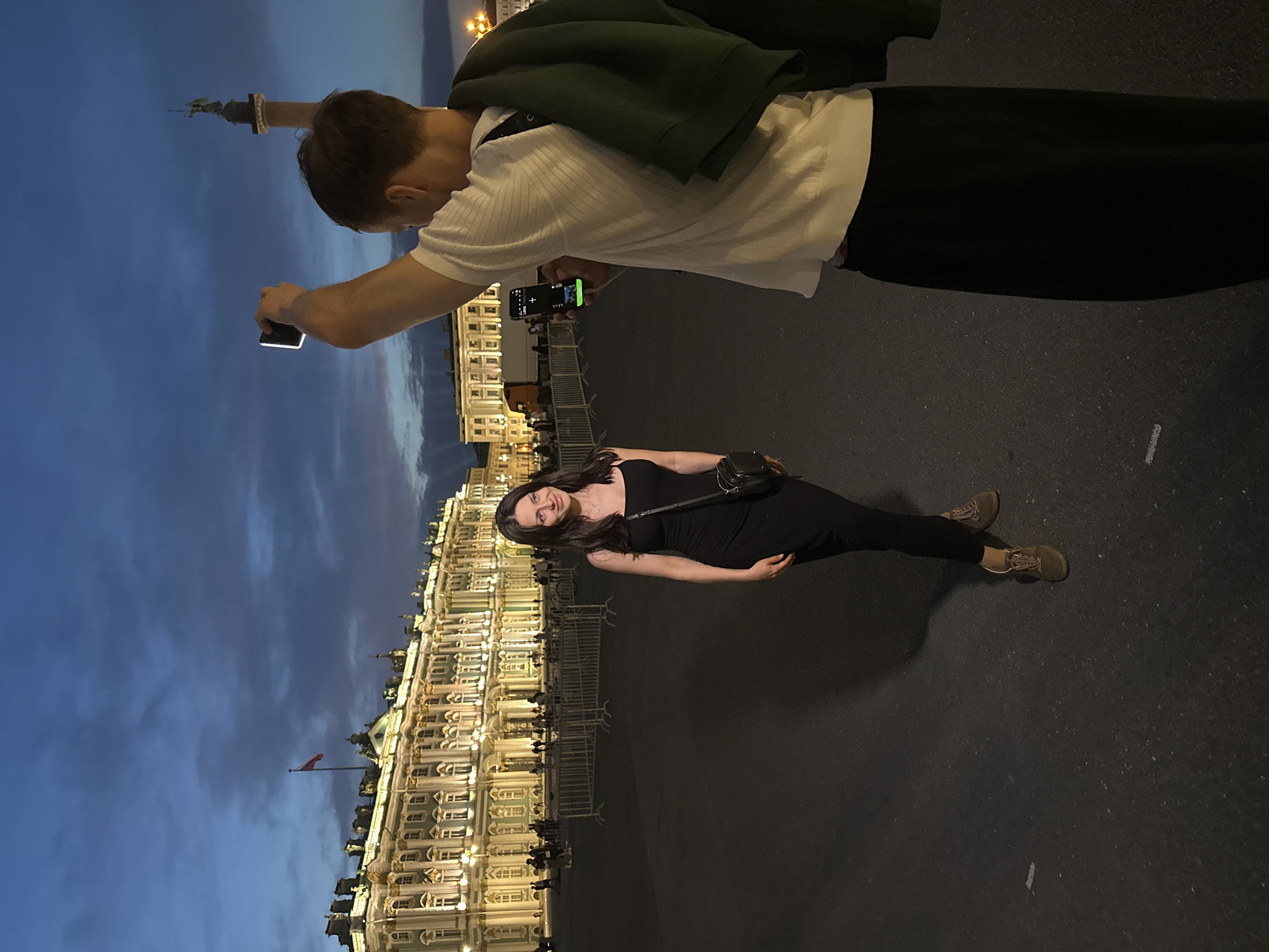 In a vast, open square at dusk, a woman in a fitted black dress poses for a photograph. She stands on the dark asphalt, her gaze slightly averted from the camera, a subtle smile gracing her lips. A black crossbody bag is slung over her shoulder. Behind her, the ornate facade of the Winter Palace, illuminated by warm lights, stretches across the horizon, its architecture hinting at historical significance. A Russian flag flutters atop a distant flagpole. To the right, a man, seen from the back, holds up a smartphone, apparently capturing the moment. He wears a white t-shirt and dark trousers, with a dark green jacket draped over his shoulder. His arm is raised, the phone's screen glowing. The sky above is a deep blue, transitioning into twilight with wisps of clouds. The scene unfolds in Palace Square, Saint Petersburg, Russia, a location recognized for its iconic historical landmarks.