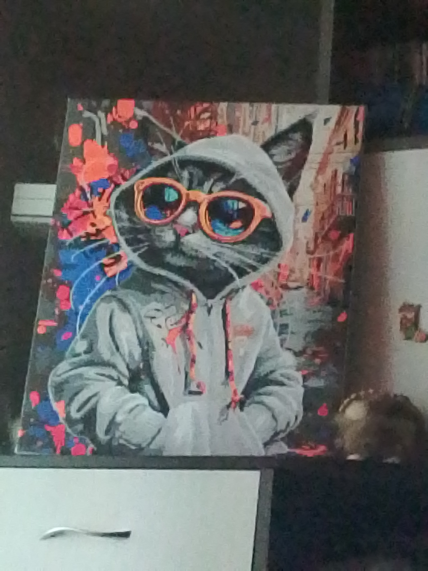 The image displays a painting of a cat wearing a hoodie and orange sunglasses. The cat is depicted in a cool, urban style, with a vibrant, abstract background that includes splashes of orange and blue. The painting is placed on a shelf or surface, with a dark, textured background behind it. To the right of the painting, there is a fuzzy, brown object that resembles a stuffed animal or decorative item. The bottom of the image shows a white drawer with a metallic handle. The overall scene appears to be indoors, possibly in a home or studio environment. There is no indication of Syasstroy, Russia, or any specific city location.