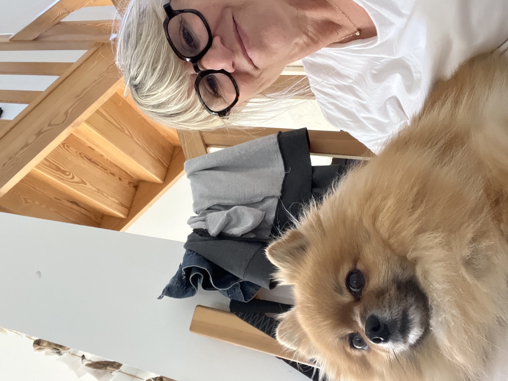 An indoor, eye-level photograph taken in Chambœuf, France, depicts an adult woman and a small, reddish-brown Pomeranian-type dog. The woman, positioned on the right, has blonde/grey hair, wears black-framed eyeglasses, a white t-shirt, and a gold necklace with a small pendant, exhibiting a subtle smile. The dog, occupying the left foreground, is fluffy with dark eyes and a black nose, gazing slightly to the left. In the background, a light-colored wooden staircase rises diagonally on the right. To the left, a white wall is visible behind a wooden furniture piece and several draped articles of clothing, including denim jeans and dark tops. The scene is brightly illuminated.