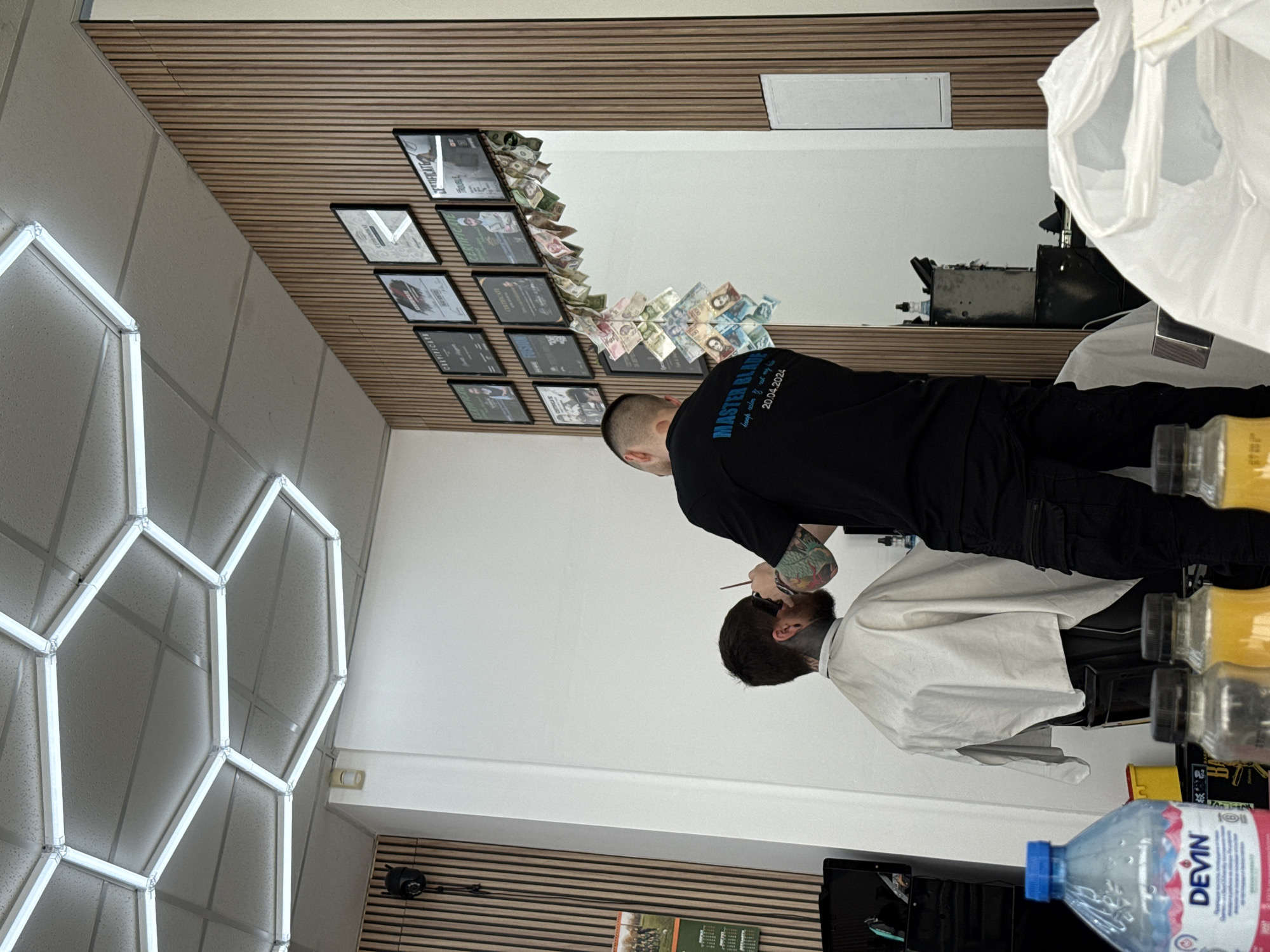 This image captures an indoor scene within a barbershop or hair salon in Sofia, Bulgaria. The central activity shows a male barber, dressed in a black t-shirt and dark trousers, meticulously cutting the hair or beard of a male client. The barber has visible tattoos on his right arm and is focused on his work, holding clippers and a comb. The client is seated, facing away from the camera, wearing a white cape.
The setting features a modern aesthetic. The wall directly behind the barber and client (reflected in the mirror) is white, while the ceiling above is made of white acoustic tiles with a hexagonal LED light fixture on the left. A prominent feature is a large mirror reflecting the shop interior. On the wall adjacent to the mirror, there are several framed items, likely certificates or photographs, arranged in a grid. Interspersed and behind these frames, numerous banknotes of various currencies, including what appear to be Bulgarian Lev, are creatively displayed, possibly stuck to the wall or mirror frame. Below this, there is a section of stylish wooden slat paneling.
On the barber's black t-shirt, text reads "MASTER BLADE", "keep calm & trust my hands", and "20.04.2024". The framed items on the wall prominently display the word "CERTIFICATE". In the foreground, on a surface near the client, several bottles are visible, including a "DEVIN" brand water bottle, indicating its Bulgarian origin. The overall ambiance suggests a professional and personalized grooming service. The time of day is not discernible due to artificial lighting.