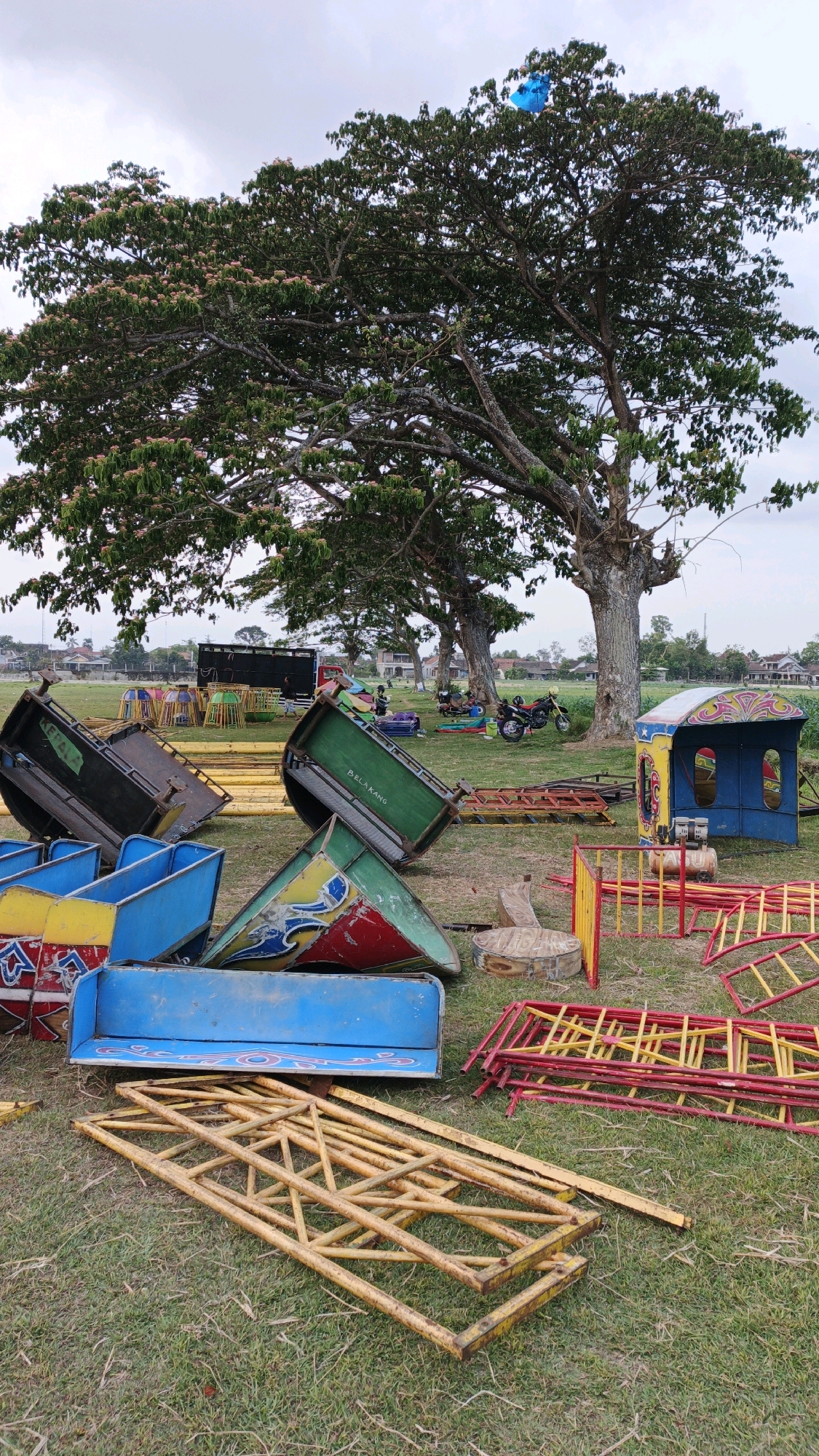 In Boyolangu, Indonesia, a collection of disassembled amusement park ride components lies scattered across a grassy field beneath a large, leafy tree. Several brightly colored ride cars, some predominantly blue with red and yellow accents, are overturned and leaning against each other. Other pieces of equipment, including what appears to be a yellow metal frame structure and red and yellow barriers, are also visible on the ground. In the background, a large truck, a motorcycle, and other structures suggest a temporary setup for an event. A bright blue balloon is caught in the upper branches of the tree. The environment appears to be an open field, possibly a park or public space, with some buildings visible in the distance. The scene conveys a sense of an amusement attraction being set up, dismantled, or temporarily stored.