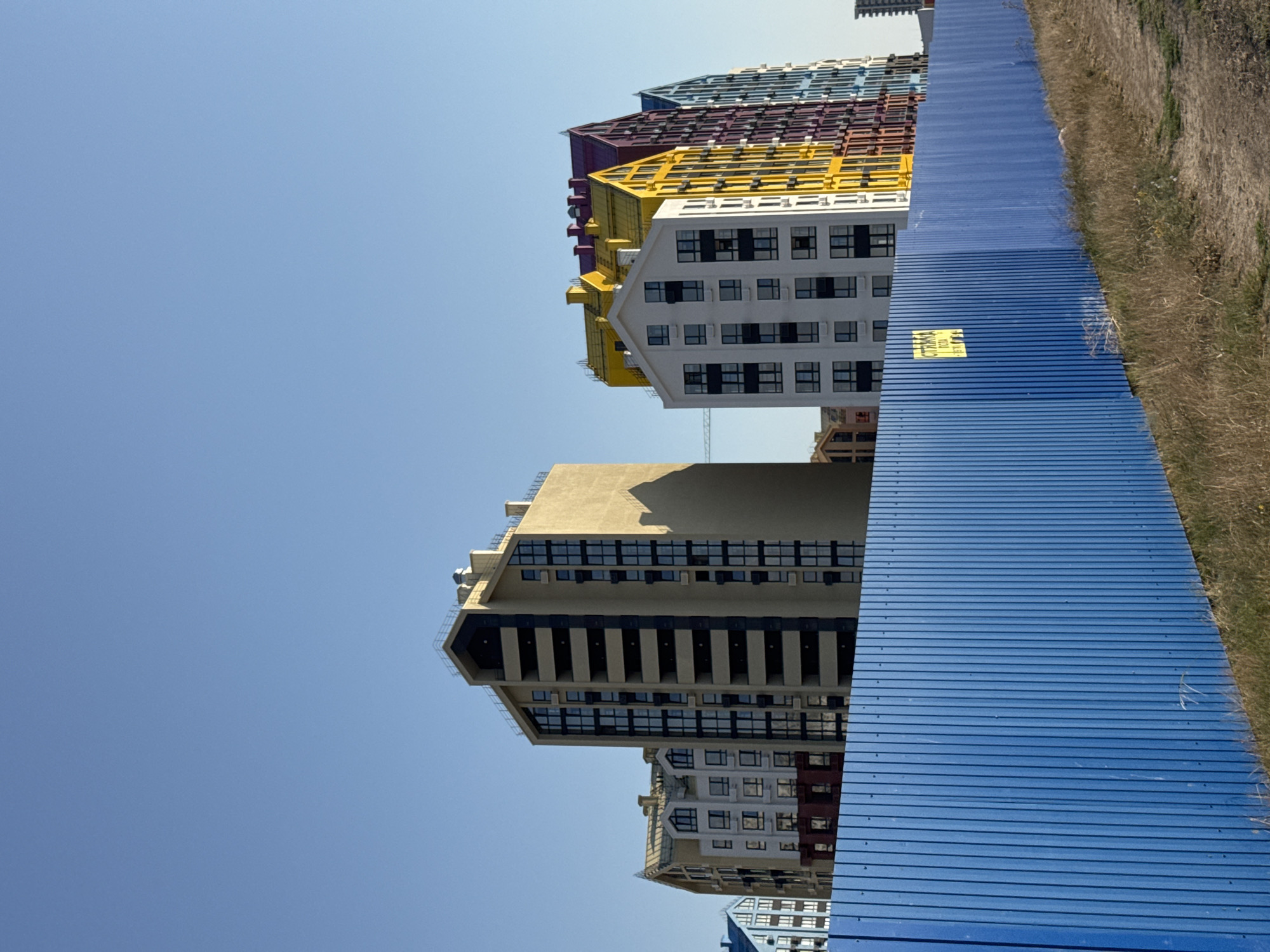 The image captures a construction site in Yevpatoriya, Ukraine, featuring a row of modern residential buildings with colorful, pitched roofs in yellow, purple, and blue, alongside a taller, beige building with dark-framed windows. A bright blue corrugated metal fence runs across the foreground, partially obscuring the construction area and the base of the buildings. Dry grass and a dirt path are visible in front of the fence. The sky is clear and blue, indicating a sunny day. No people are present in the frame.