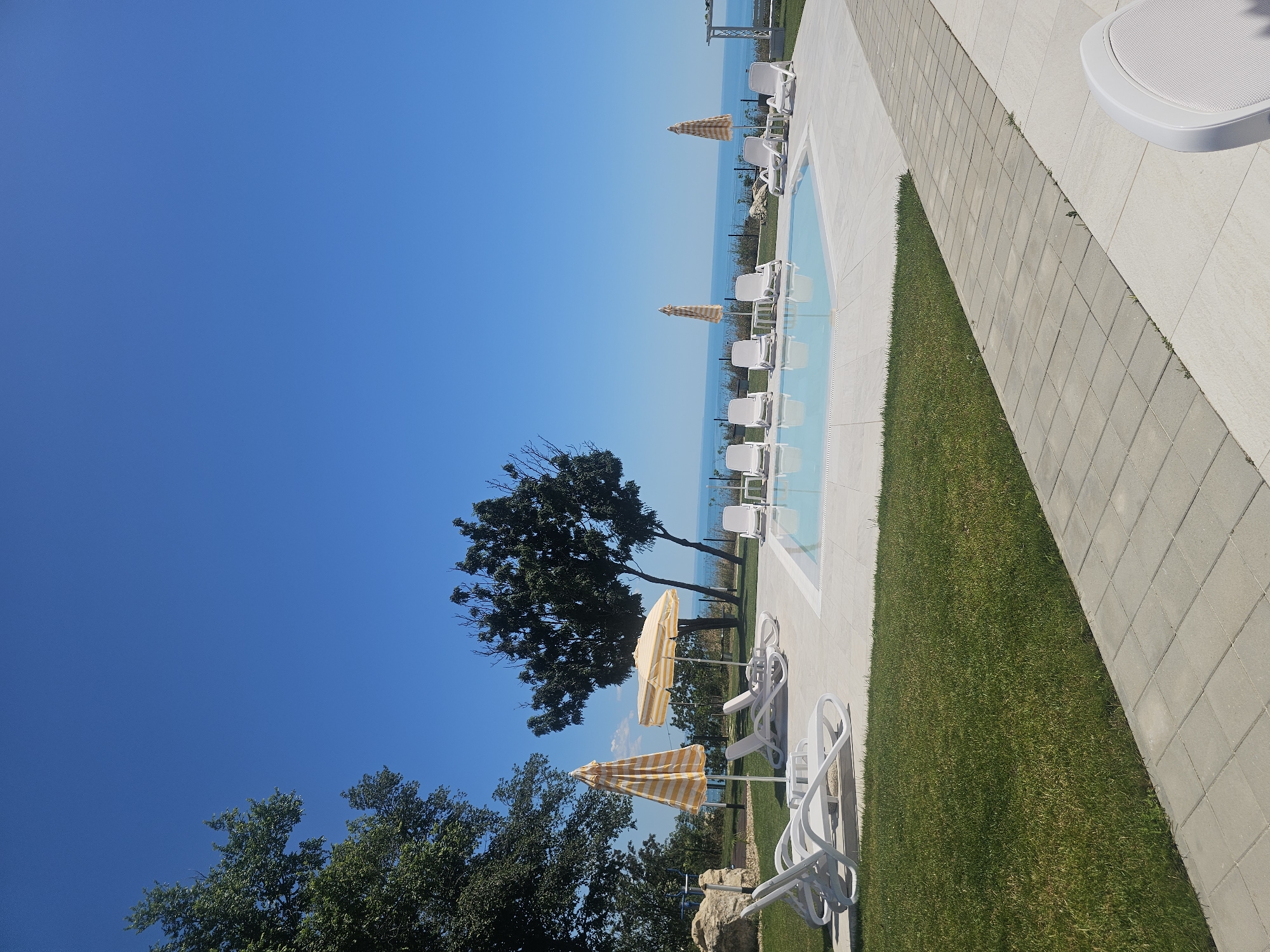 The image depicts an outdoor swimming pool area with a clear blue sky and the ocean in the background. The pool is irregularly shaped and filled with clear water, with white tiles lining its edges. Around the pool, there are several white lounge chairs and some striped yellow and white umbrellas providing shade. The area is bordered by a neatly mowed green lawn and a paved walkway made of gray square tiles. In the distance, beyond the pool and lawn, is a dark fence separating the property from a sloping grassy area leading down towards the sea. Trees with green foliage are visible on the left and in the middle ground, casting shadows. The horizon shows a clear divide between the blue of the sea and the sky. The overall scene is tranquil and suggests a resort or private residence. There are no people visible in the image.